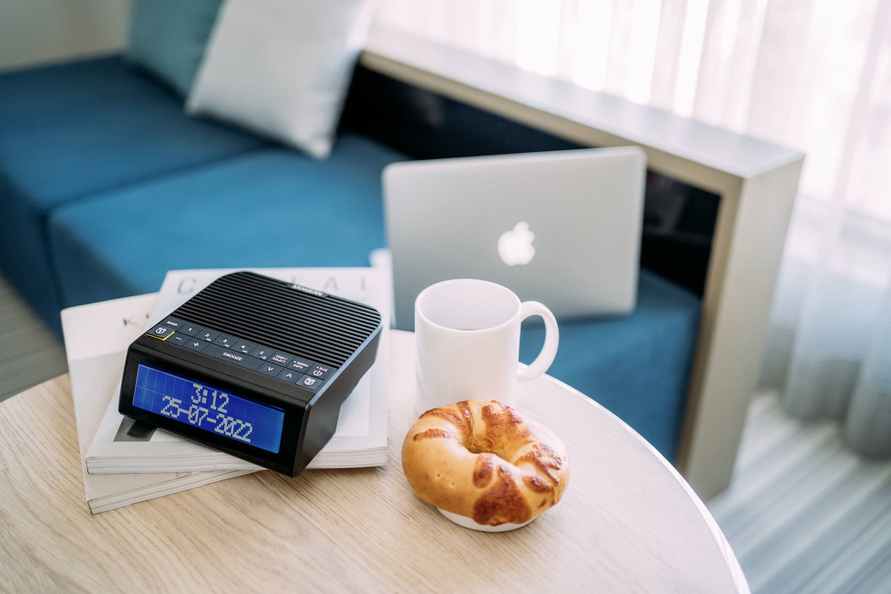 Ein Tisch mit Kaffee, Croissant, digitalem Wecker und Laptop neben Sofa. Uhrzeit auf Wecker: 9:12.  Gemütliche Arbeitsecke im Wohnzimmer.