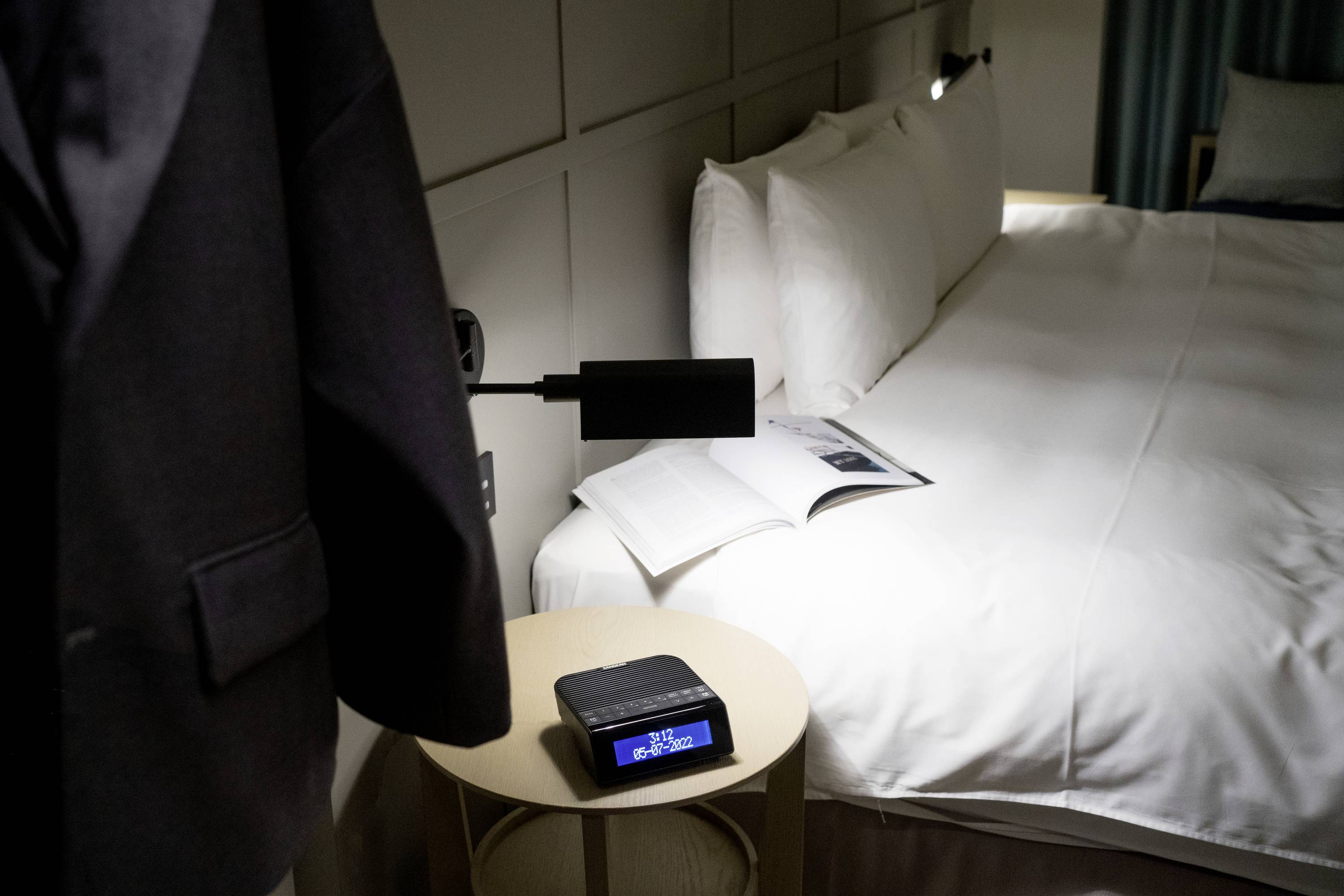 Ein Hotelzimmer mit einem Bett, darauf ein aufgeschlagenes Buch. Daneben ein Tisch mit einem Digitalwecker, auf dem die Uhrzeit angezeigt wird. Ein Sakko hängt am Stuhl.