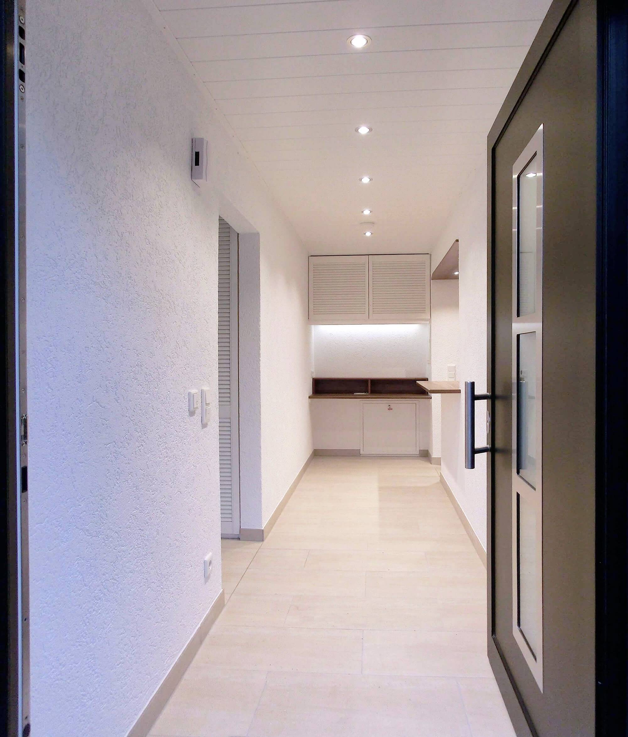 Ein moderner Flur mit hellem Boden und weißen Wänden, führt zu einem offenen Raum mit heller Holzoberfläche und Einbauschränken.