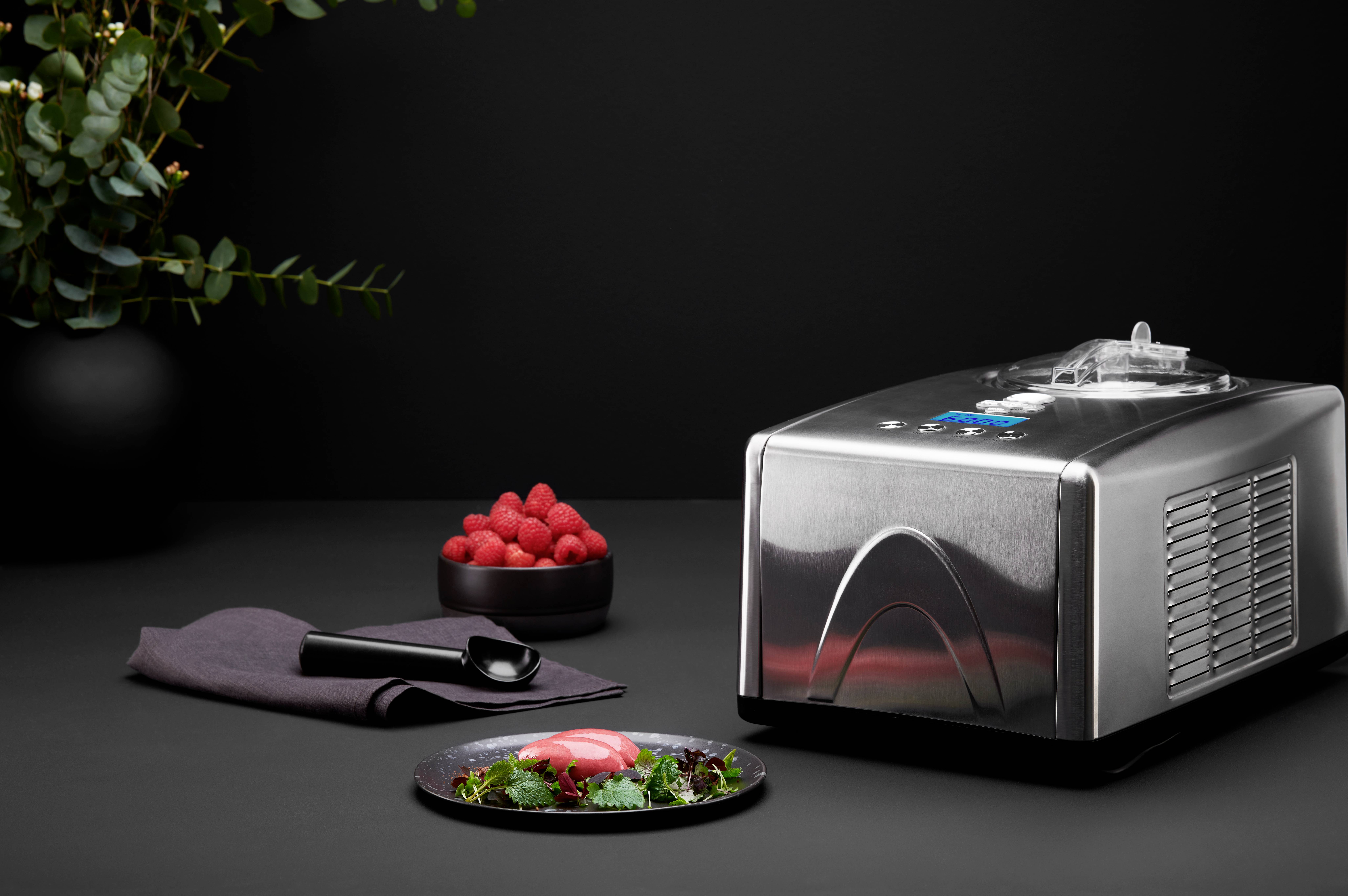Eine Chrom-Eismaschine neben einem Teller mit Dessert, einer Schale Himbeeren und einem schwarzen Löffel, vor dunklem Hintergrund.
