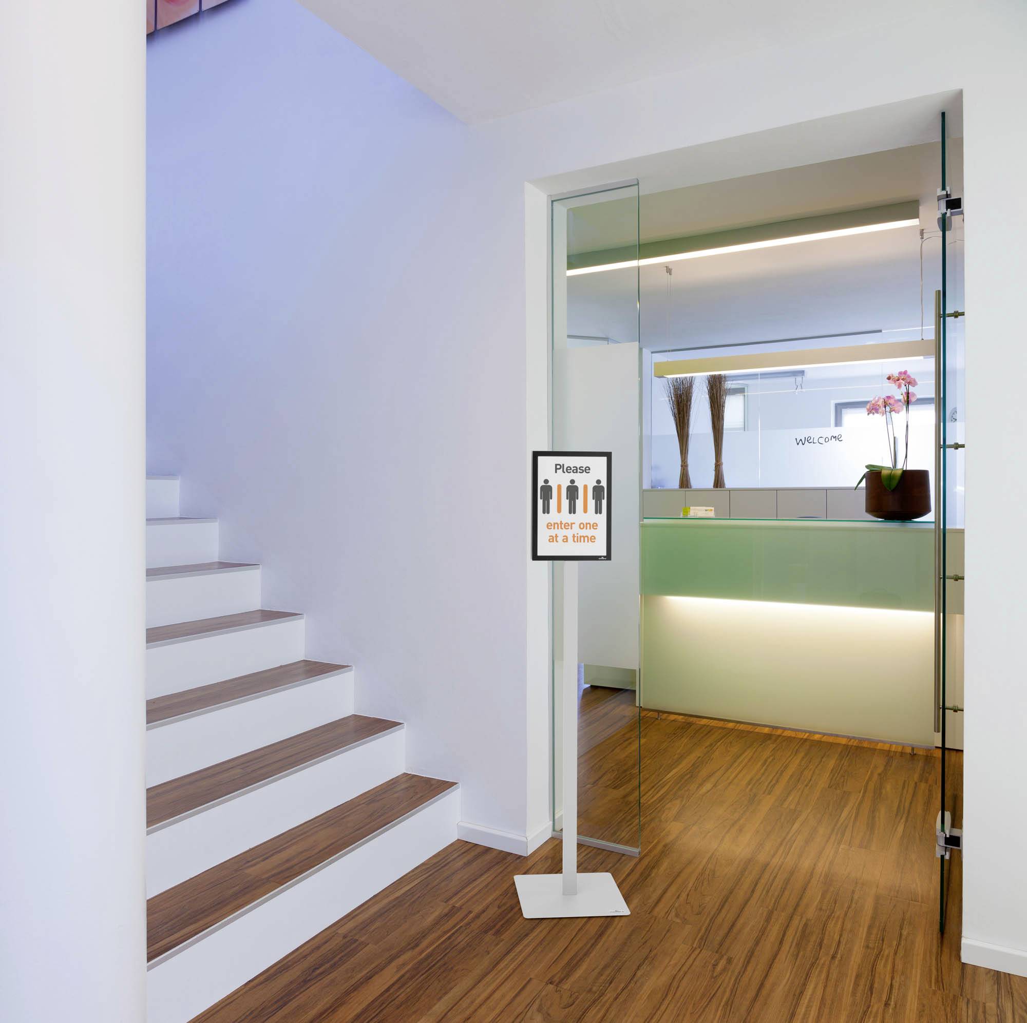 Eine offenstehende Glastür neben einer Treppe, mit einem Schild, das 'Bitte Abstand halten' zeigt. Dahinter ein moderner Empfangsbereich.