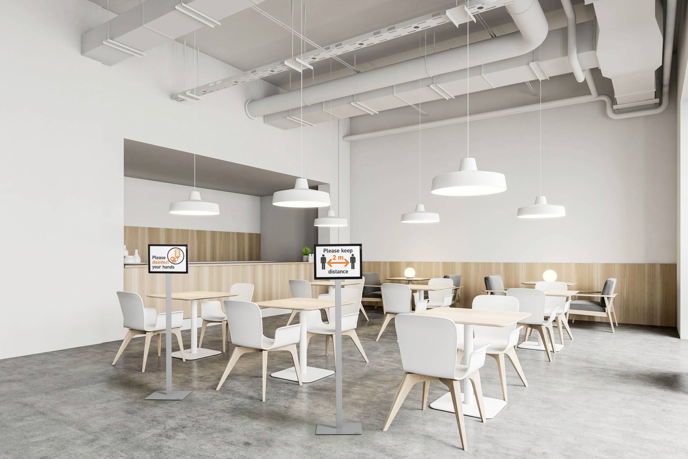 Ein moderner Speisesaal mit weißen Tischen und Stühlen, hellen Hängelampen und neutralem Dekor. Zwei Bildschirme zeigen Sicherheitsinformationen.