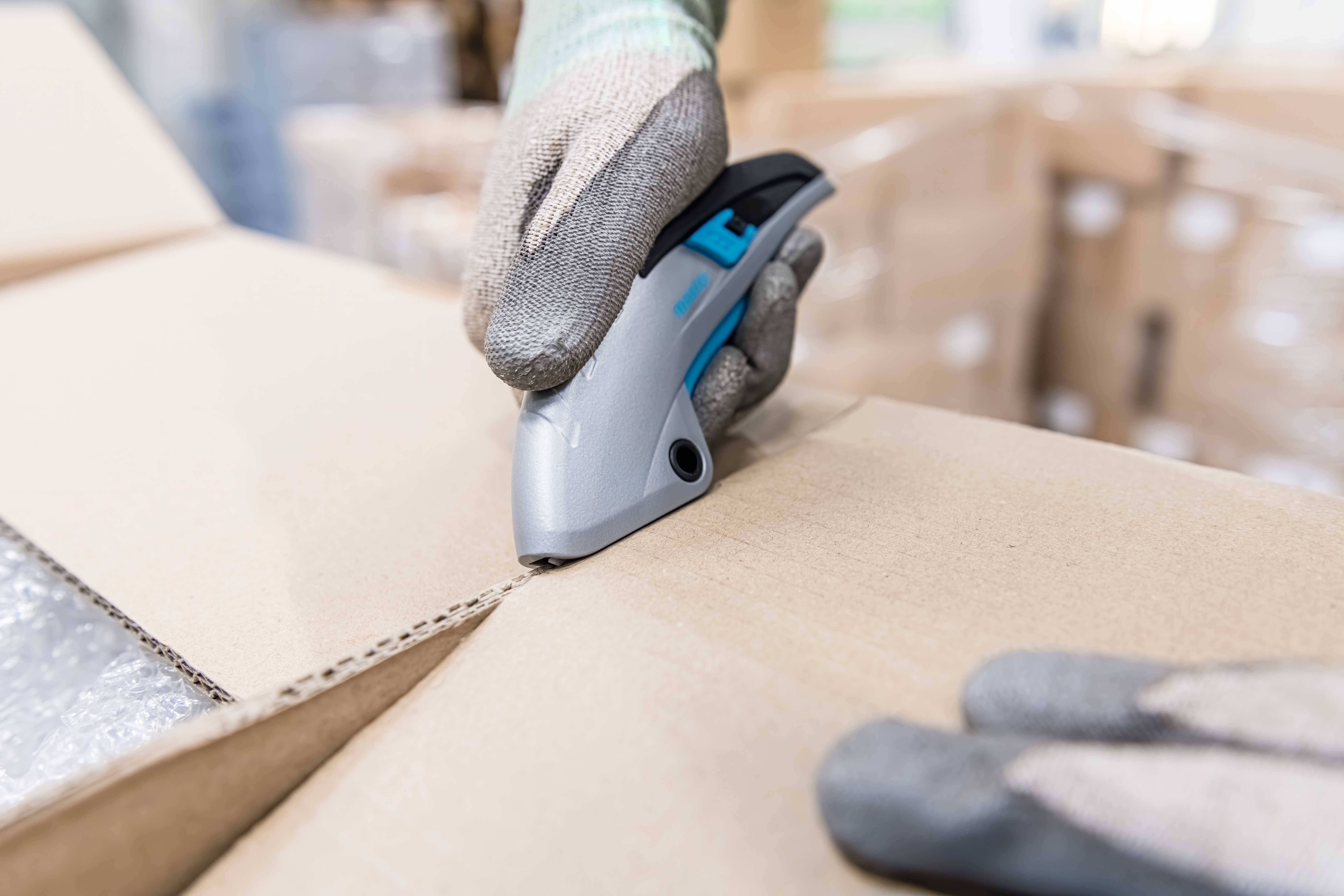 Eine Person schneidet mit einem Cutter-Messer eine Pappschachtel auf. Im Hintergrund sind unscharf Kartons zu sehen.