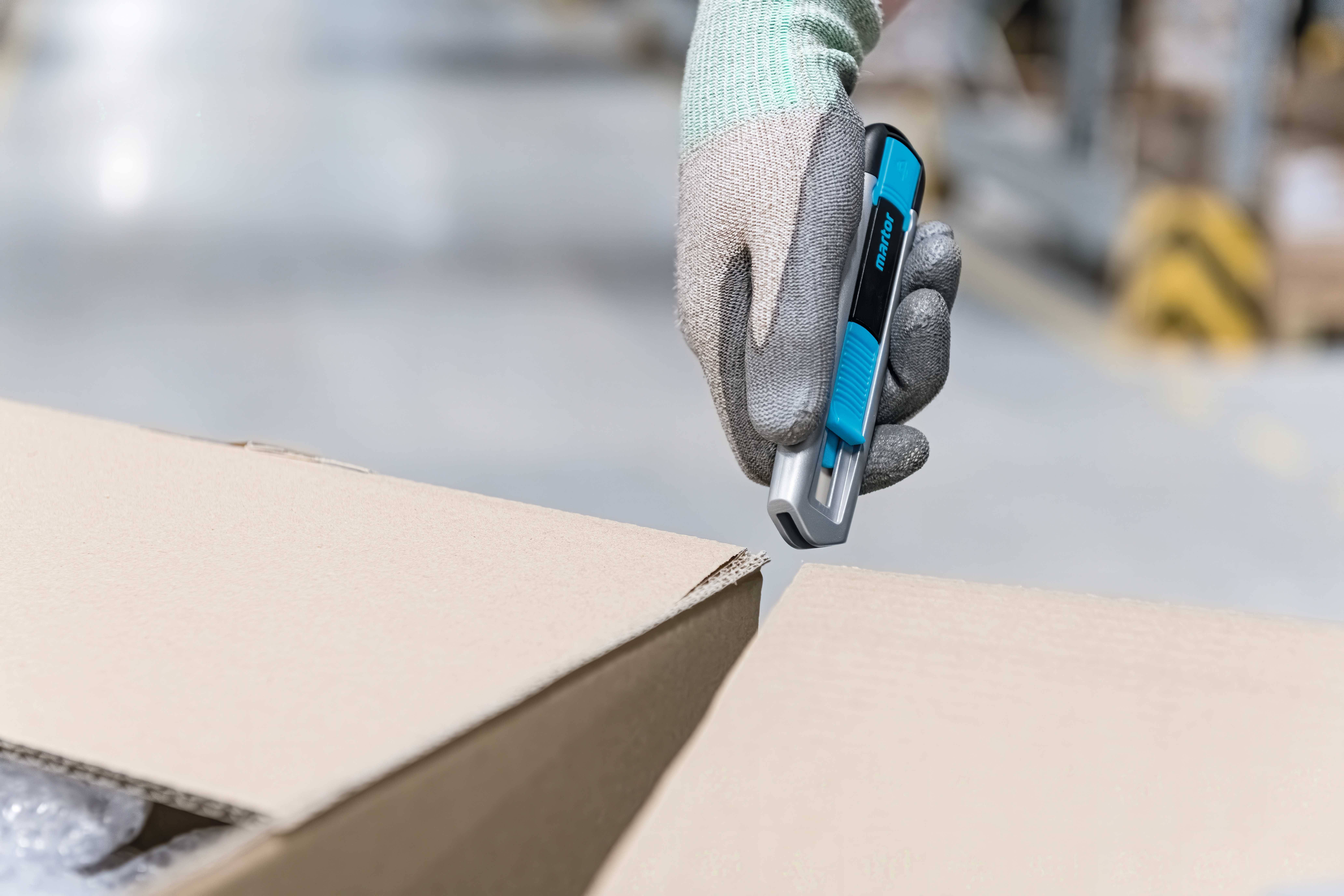 Eine behandschuhte Hand hält ein blaues Messer zum Öffnen eines Pappkartons in einer Lagerhalle.