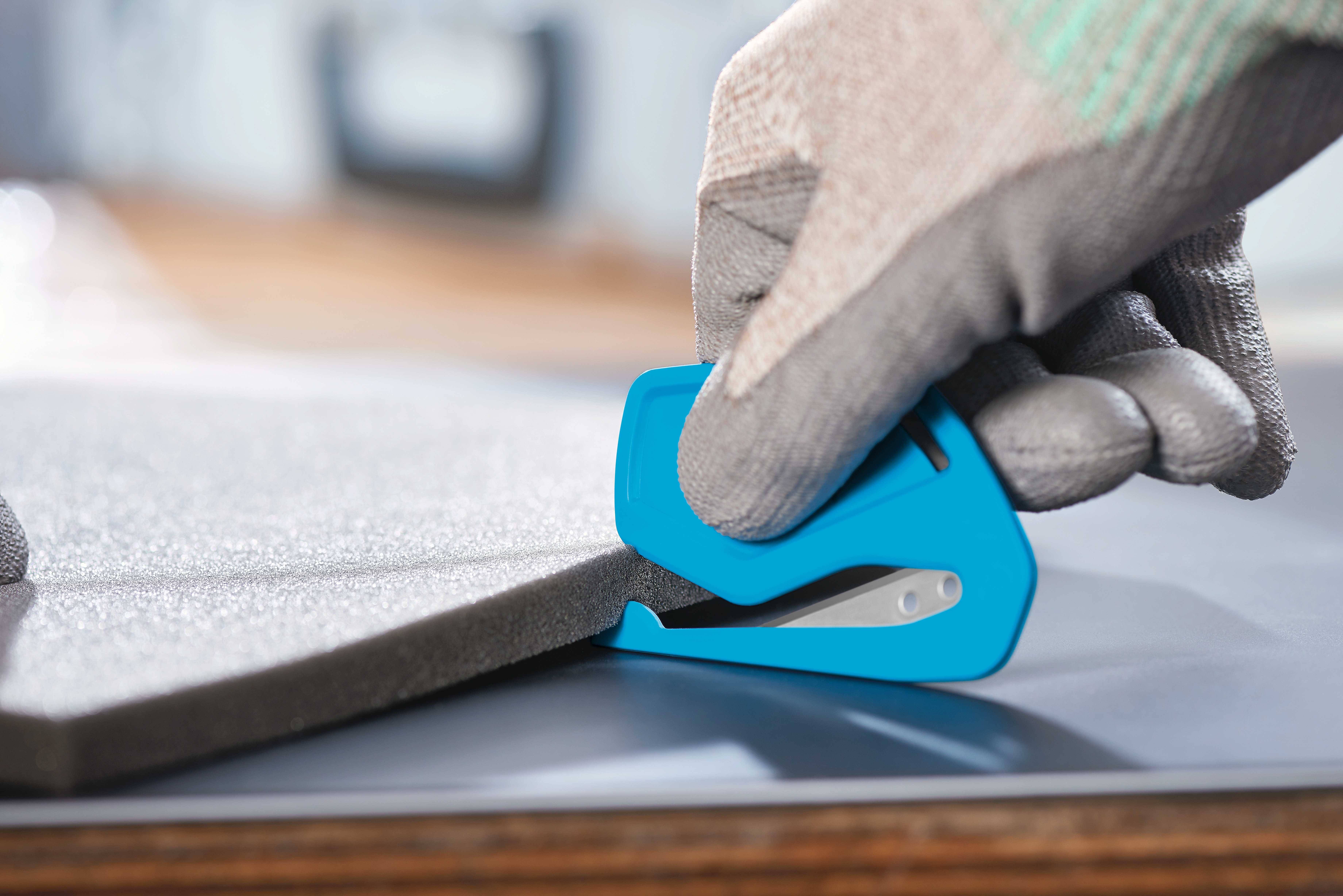 Eine Person schneidet mit einem blauen Safety Cutter ein Stück grauen Schaumstoff auf einem Tisch.