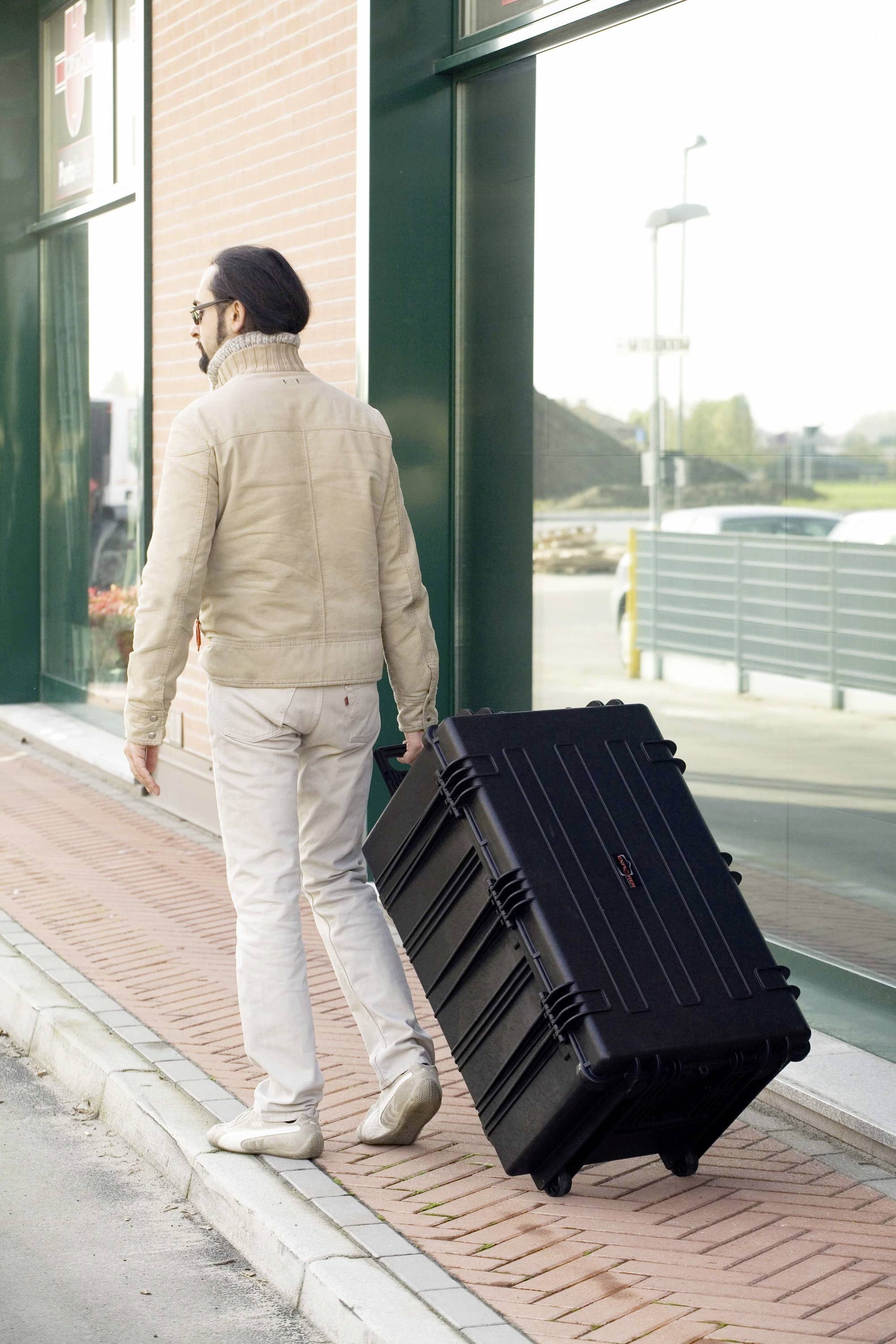 Ein Mann in heller Kleidung zieht einen großen, schwarzen Koffer auf einem Gehweg entlang eines Gebäudes.
