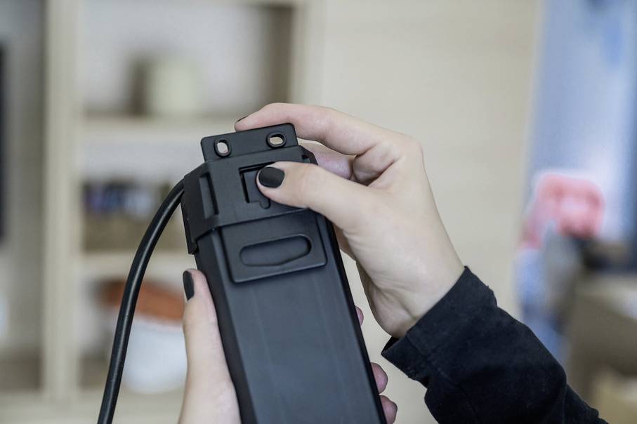 Eine Person hält ein schwarzes Gerät mit einem Kabel, das eine Art Joystick oder Bedienelement hat. Im Hintergrund ein Regal.