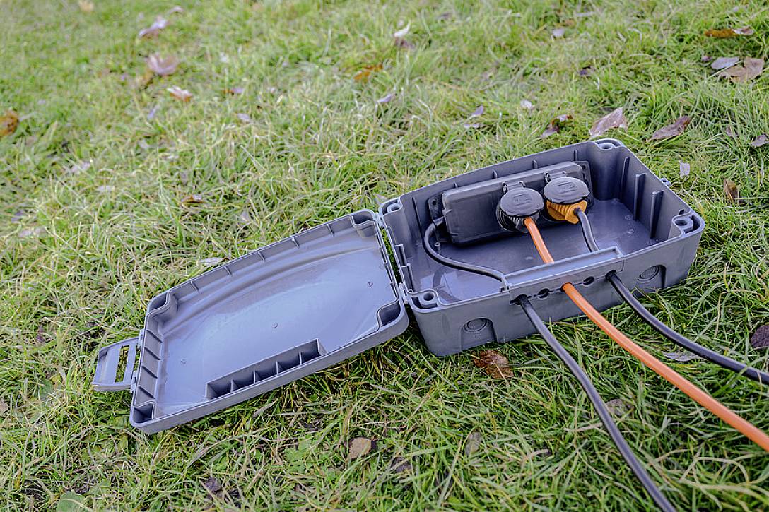 Eine offene graue Outdoor-Steckdose auf Rasen zeigt zwei eingesteckte orangefarbene Kabel. Blätter liegen verstreut auf dem Gras.