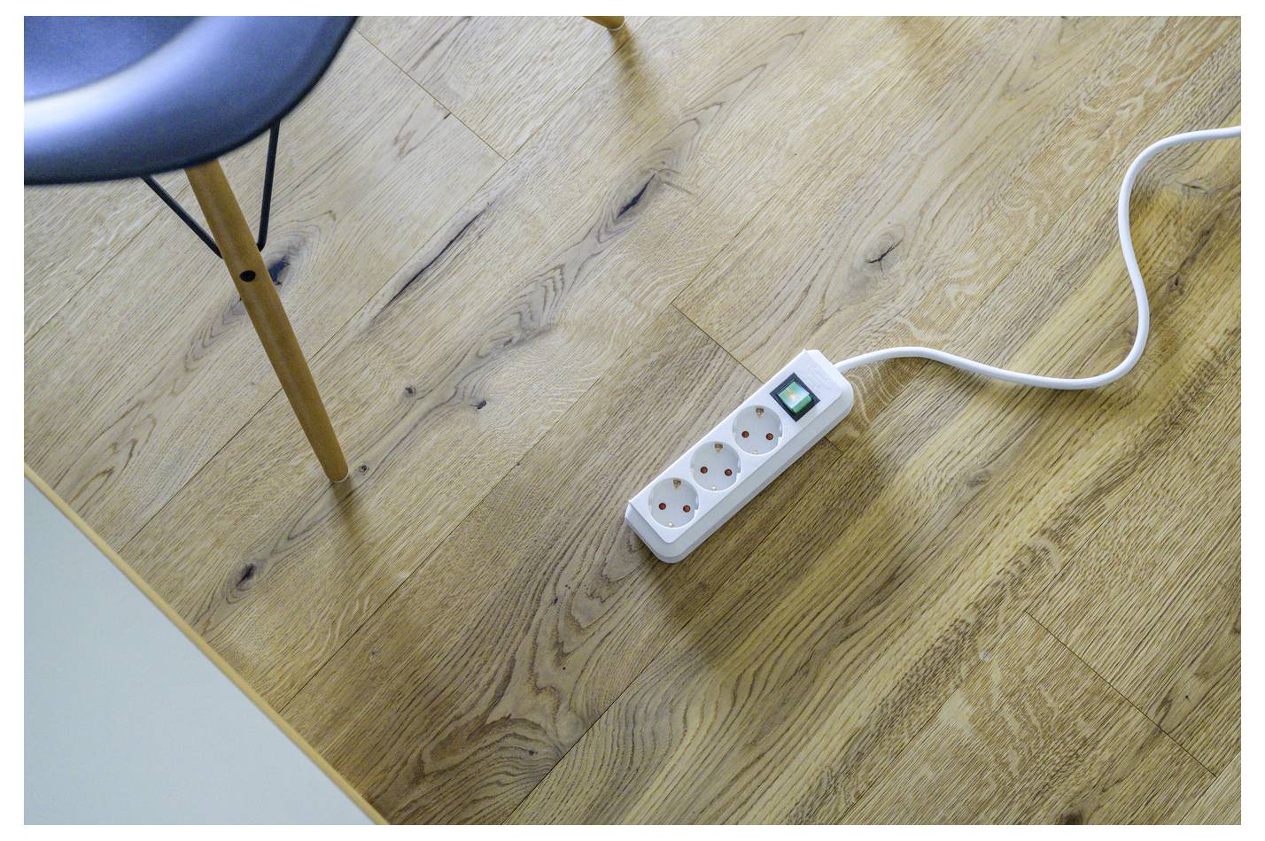 Eine weiße Steckdosenleiste mit drei Steckdosen und einem grünen Schalter liegt auf einem Holzboden neben einem Stuhlbein, während ihr Kabel aus dem Bild verläuft.