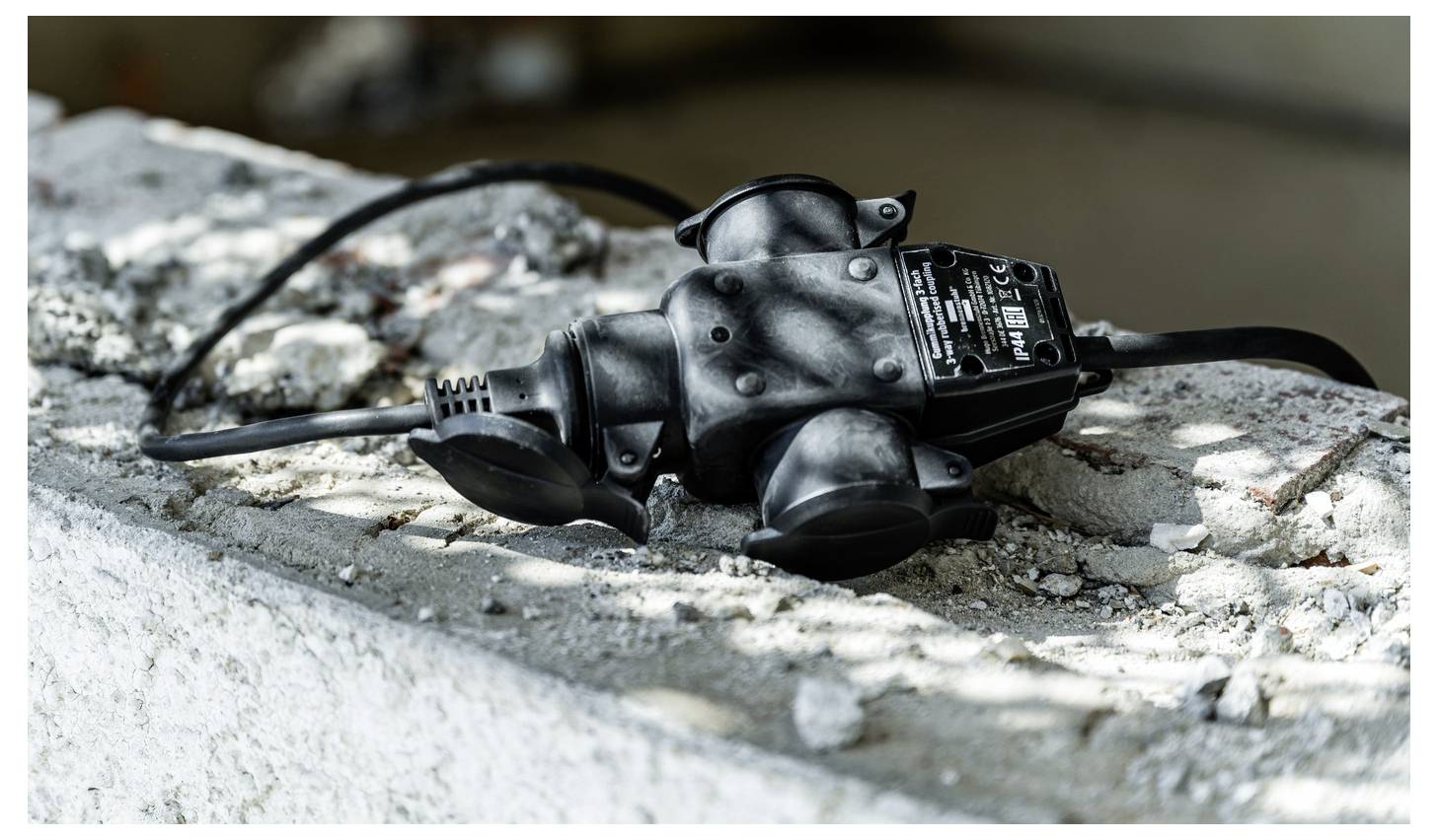 Schwarzer Stromsteckeradapter liegt auf einer rauen Betonoberfläche im Freien, mit einem angeschlossenen Kabel und einem sichtbaren Etikett oben darauf.