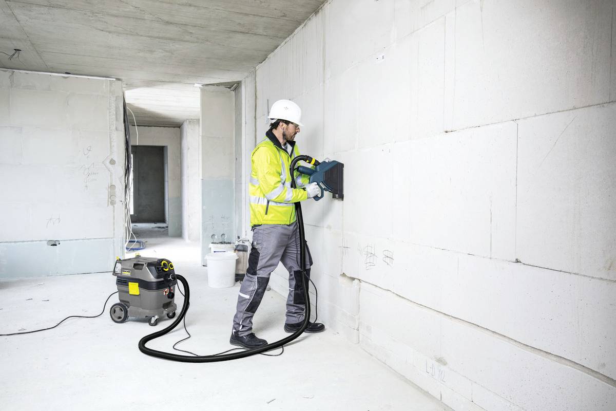 Ein Bauarbeiter in Arbeitskleidung und Helm verwendet ein elektrisches Werkzeug, um eine Wand in einem unfertigen Raum zu bearbeiten.