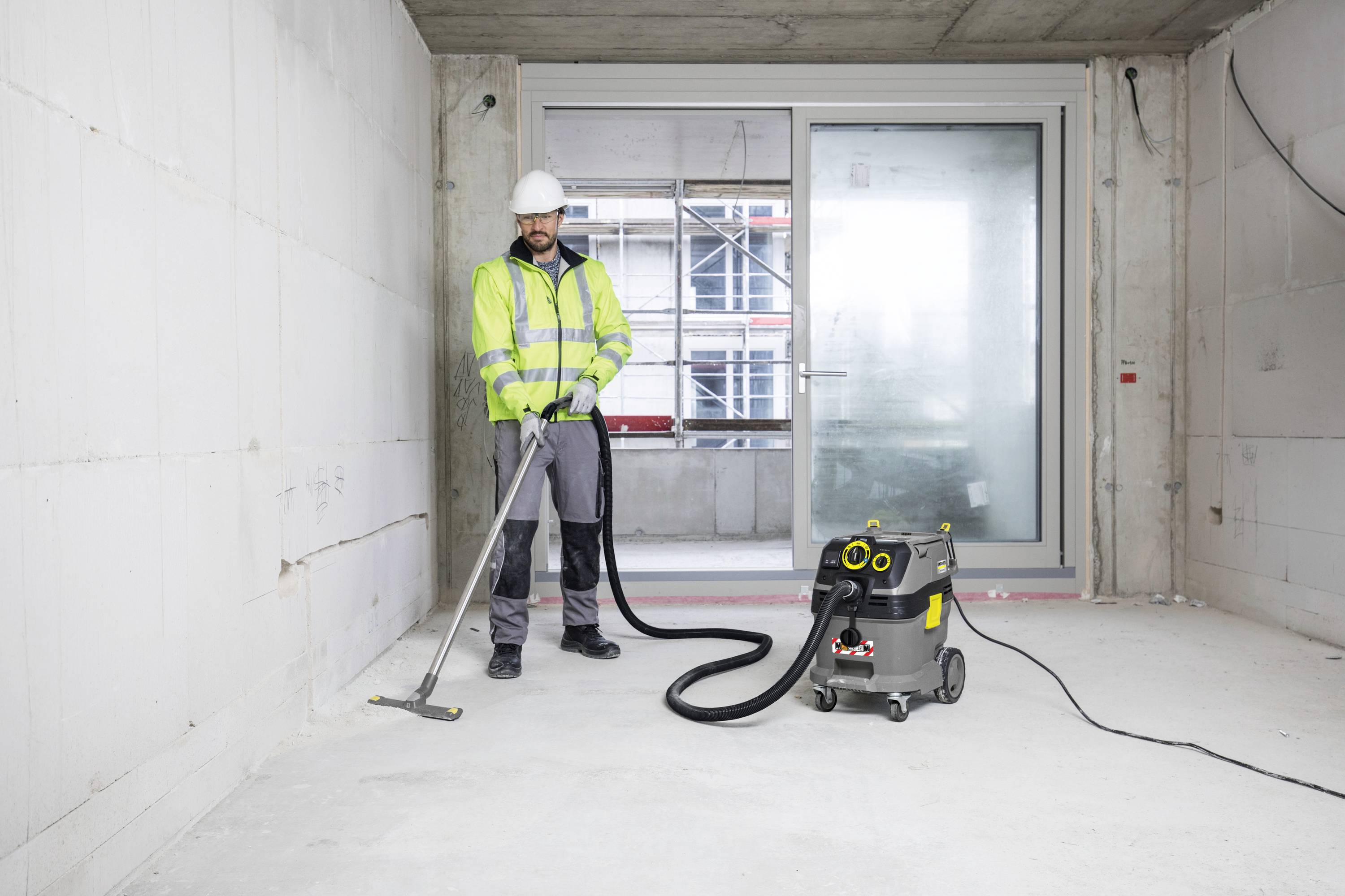 Ein Bauarbeiter in Schutzkleidung reinigt einen leeren Raum mit einem Industriestaubsauger. Große Fenster im Hintergrund.