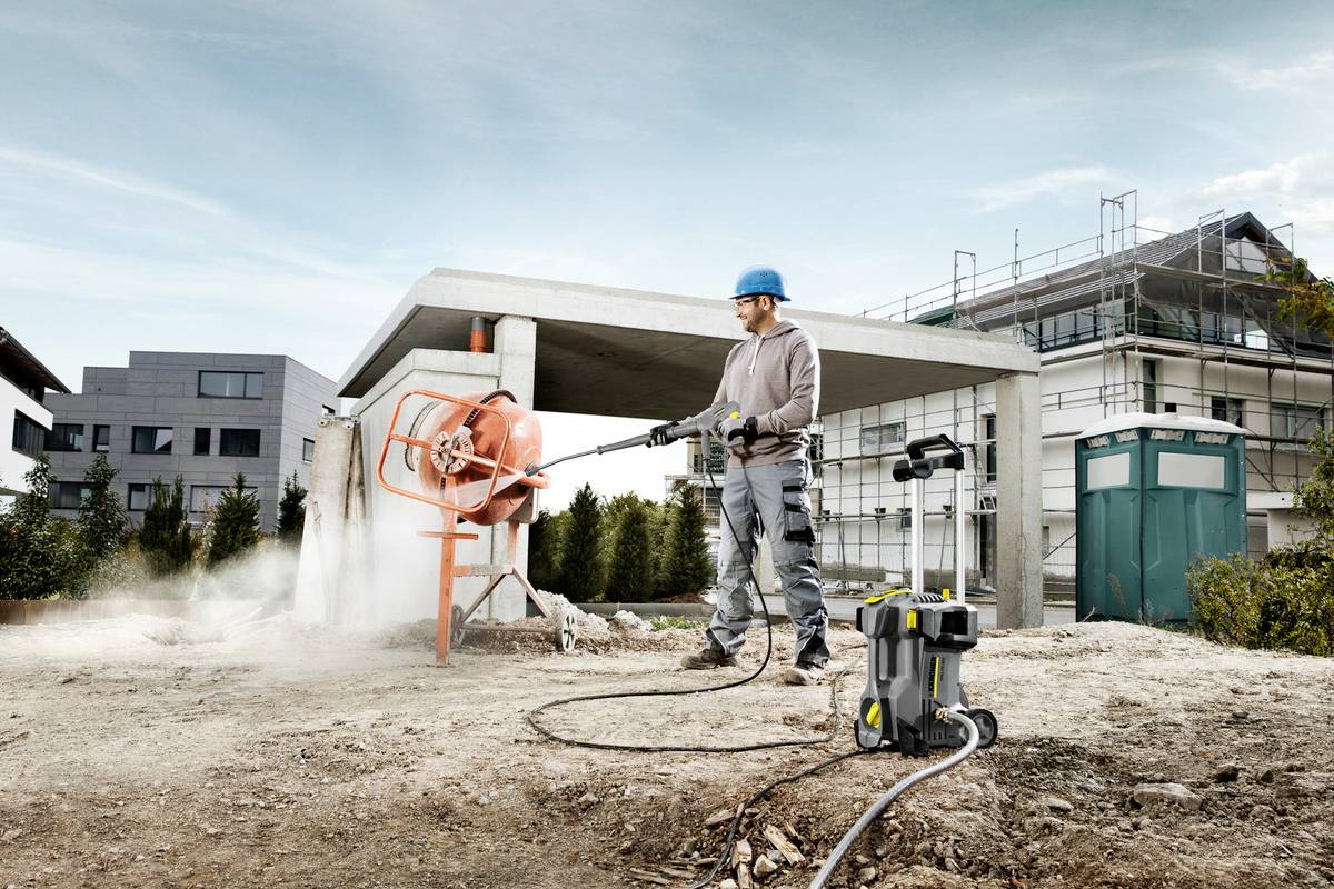 Ein Bauarbeiter mit Helm spritzt Wasser auf einen Zementmischer auf einer Baustelle. Im Hintergrund sind Gebäude und ein Gerüst sichtbar.