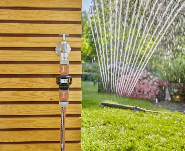 Ein Gartenschlauch, der an einer Holzwand mit einer Zeitschaltuhr befestigt ist; im Hintergrund bewässert ein Sprinkler einen grünen Rasen und Blumensträucher.