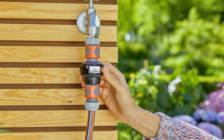 Eine Hand stellt einen digitalen Wasserzeitschalter ein, der an einem Gartenschlauch an einer Holzwand im Freien befestigt ist, umgeben von Grünpflanzen und Blumen.