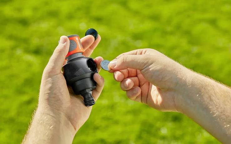 Eine Person steckt eine Münze in einen Wasserflussregler, den sie in der Hand hält, vor einem Hintergrund mit Gras.