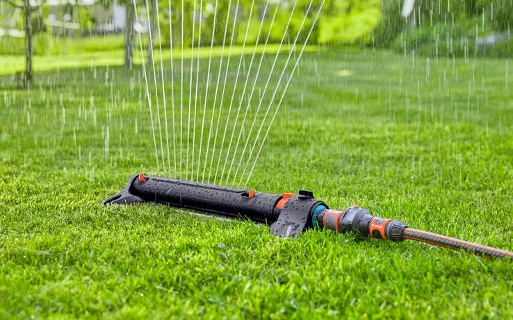 Eine Person befestigt einen orangenen und grauen Schlauchverbinder an einem Metallhahn im Freien, mit einem verschwommenen grünen Gartenhintergrund.