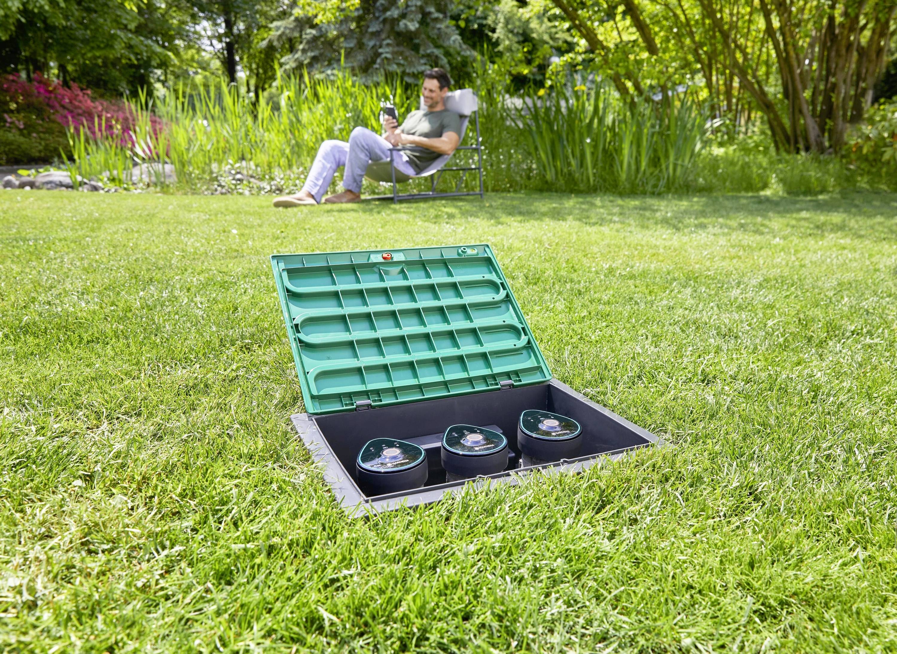 Ein Mann sitzt entspannt im Gartenstuhl im Hintergrund. Im Vordergrund ist ein offener, grüner Kasten mit drei schwarzen Behältern im Gras.