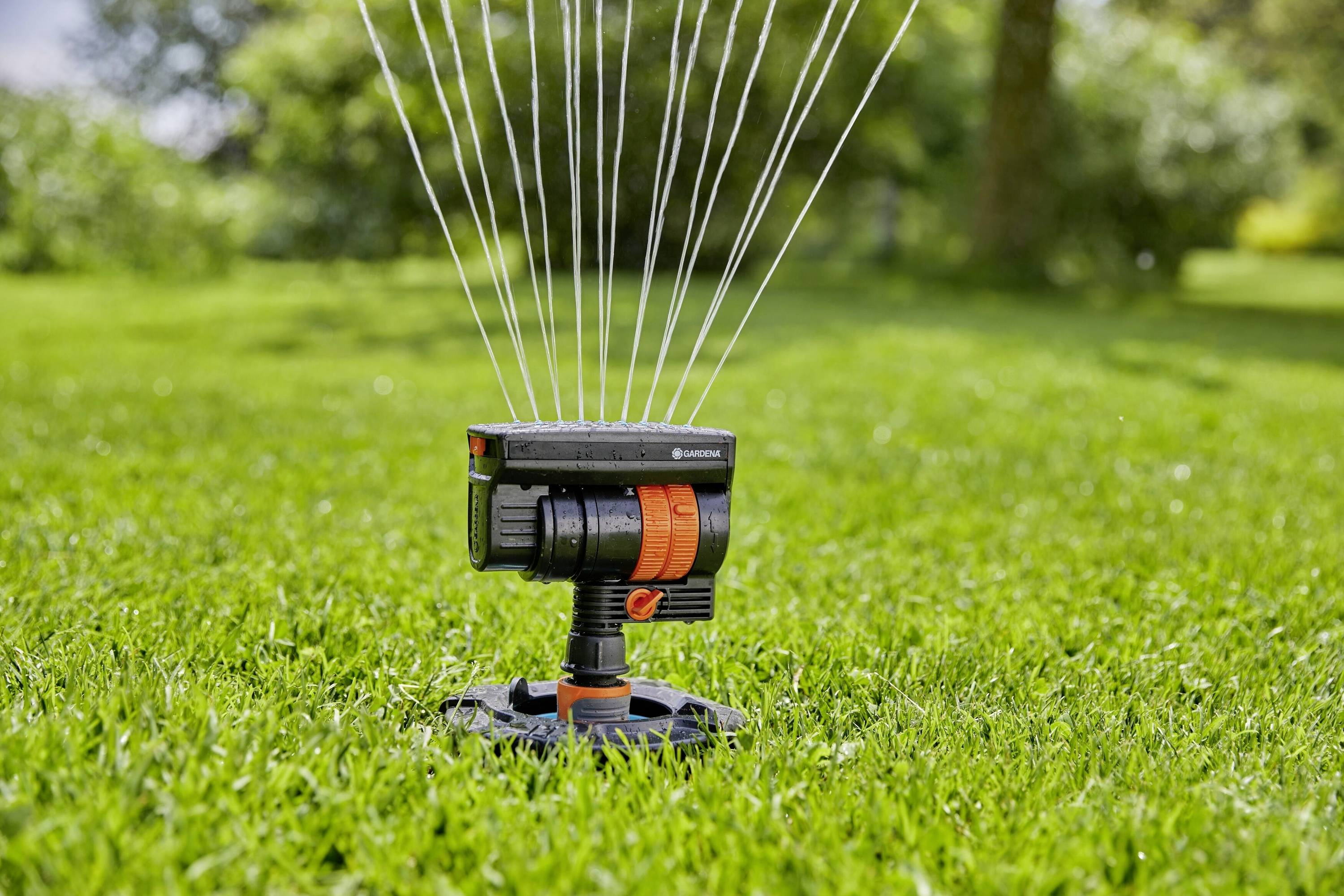 Ein Rasensprinkler auf grünem Gras, der mehrere Wasserstrahlen in die Luft befördert, um die Umgebung zu bewässern. Im Hintergrund sind Bäume sichtbar.