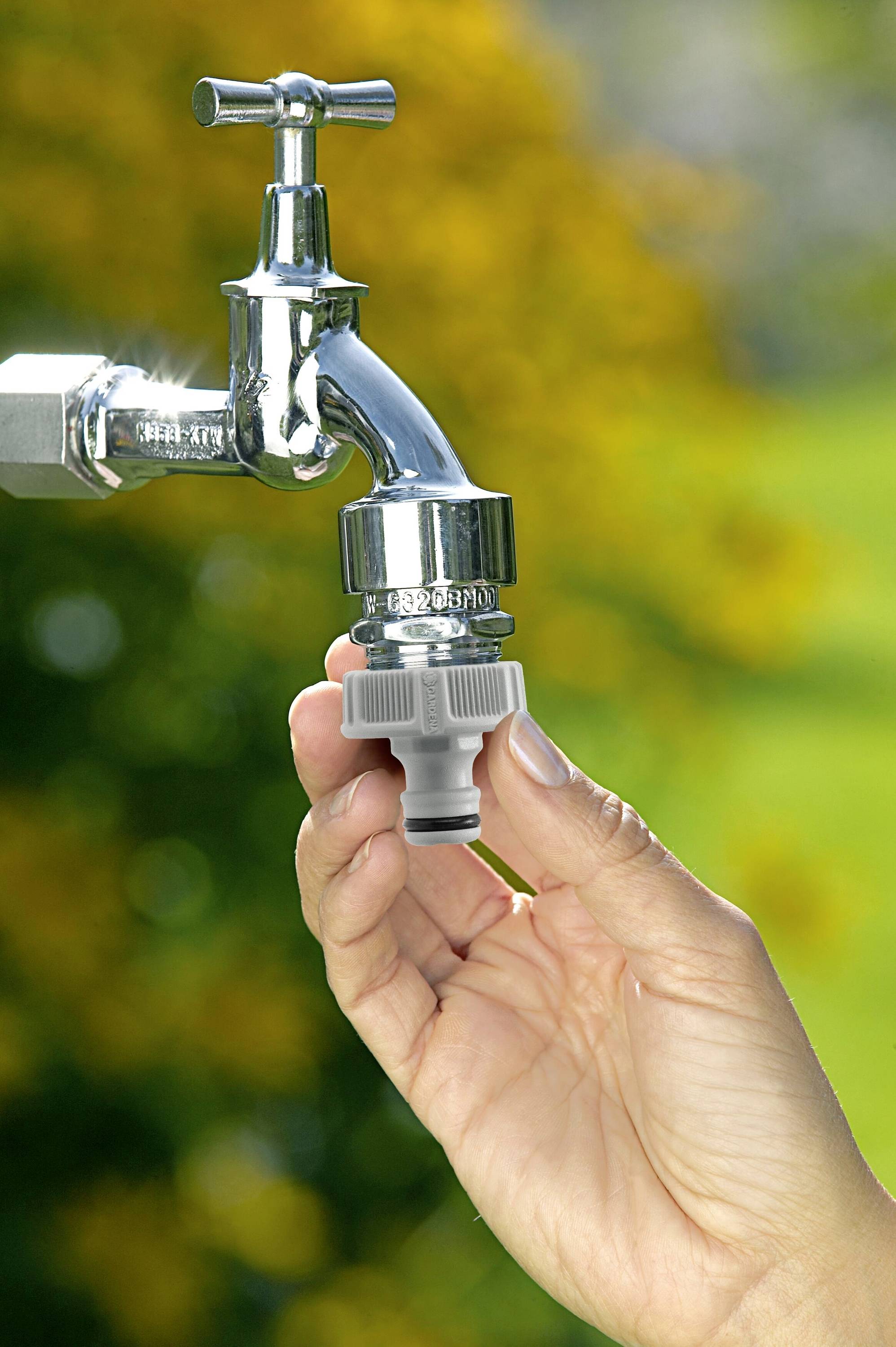 Eine Hand hält einen Wasserhahnadapter unter einen geschlossenen Außenwasserhahn vor einem Hintergrund aus grünen Pflanzen.