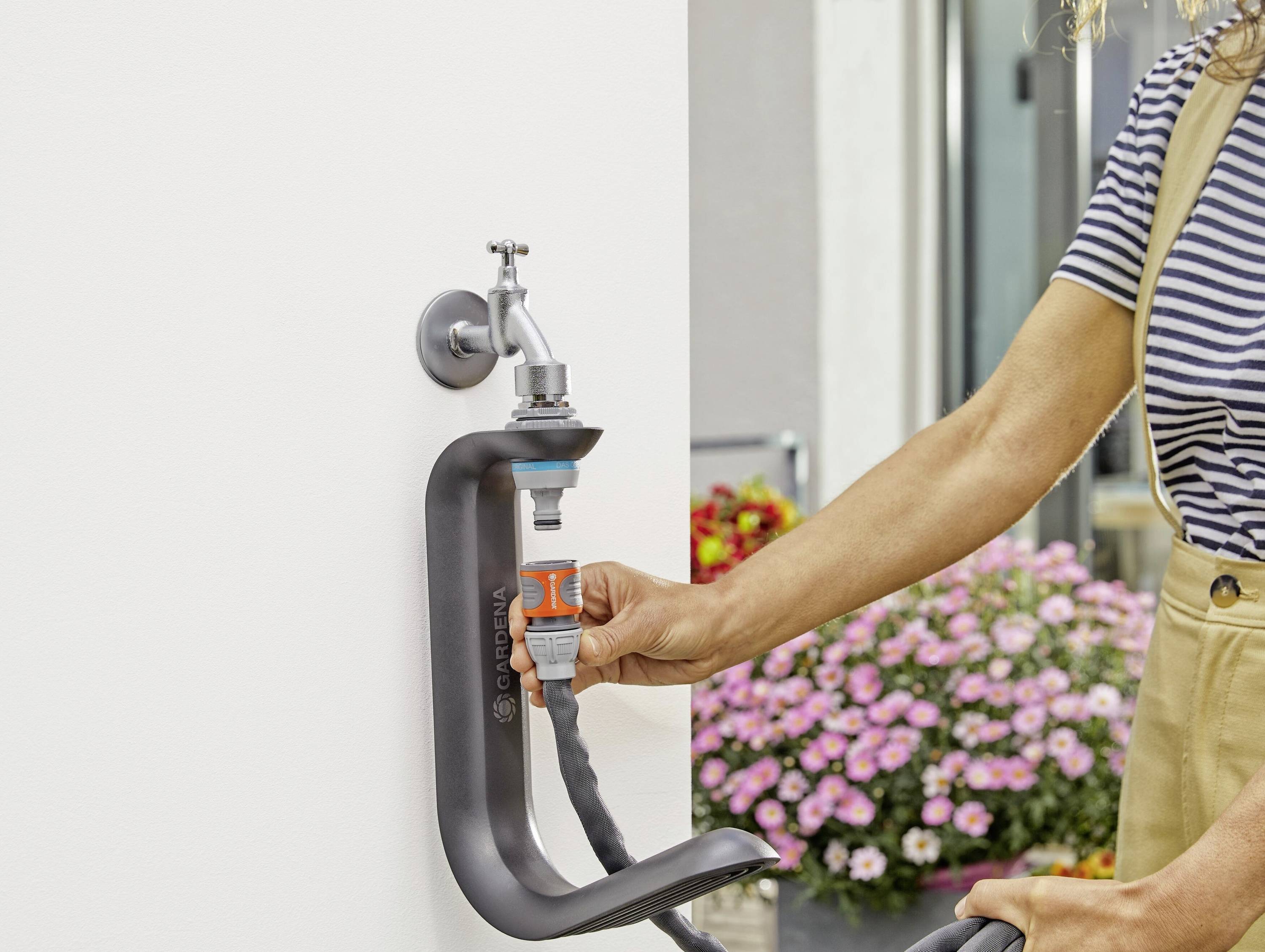 Eine Person schließt einen Gartenschlauch an einen Außenwasserhahn an. Im Hintergrund blühen rosa Blumen in einem Pflanzgefäß.