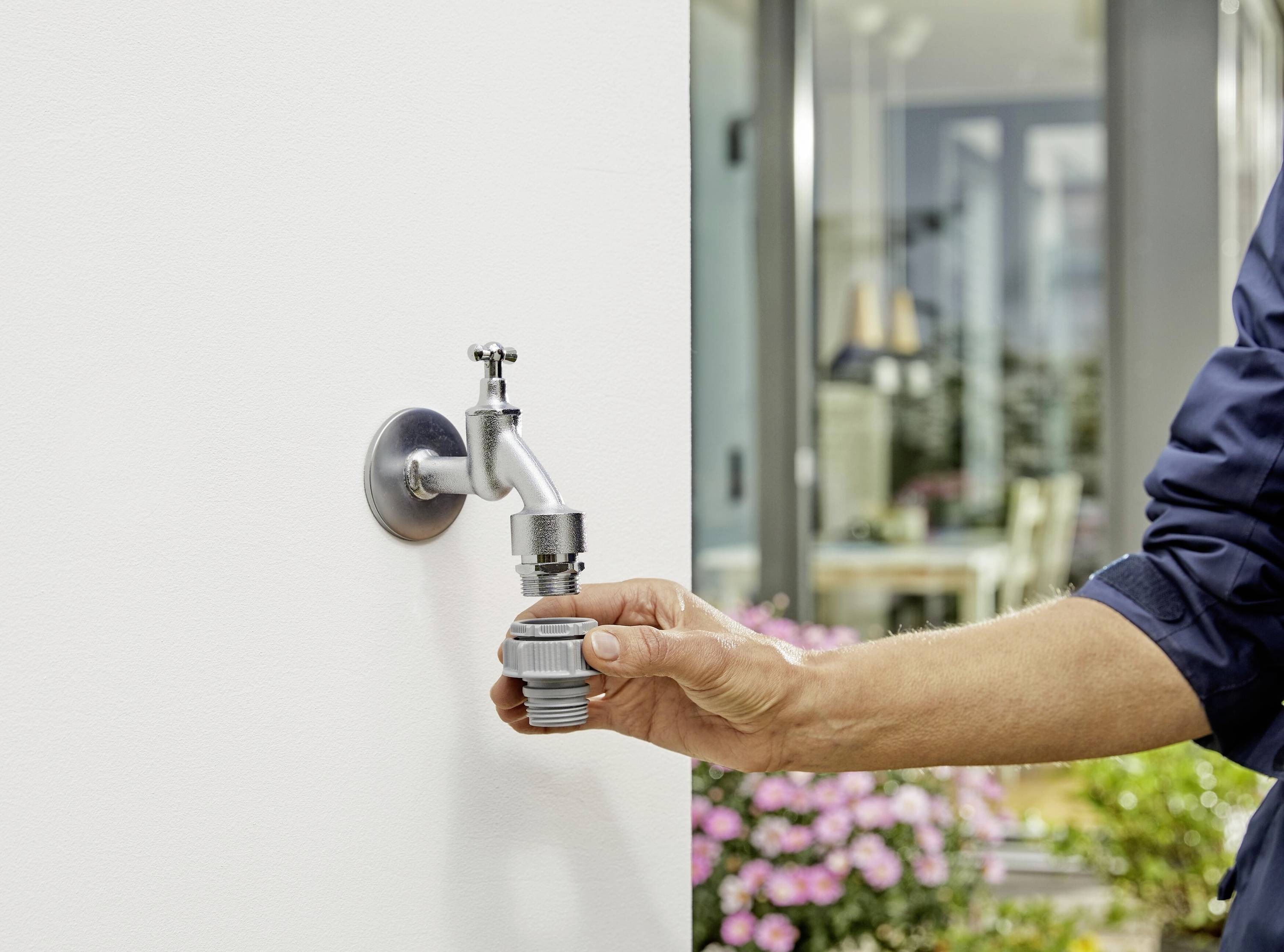 Eine Person schließt einen Gartenschlauch an einen Außenwasserhahn an einer weißen Wand an, mit einem Haus und einem Blumenbeet im Hintergrund.