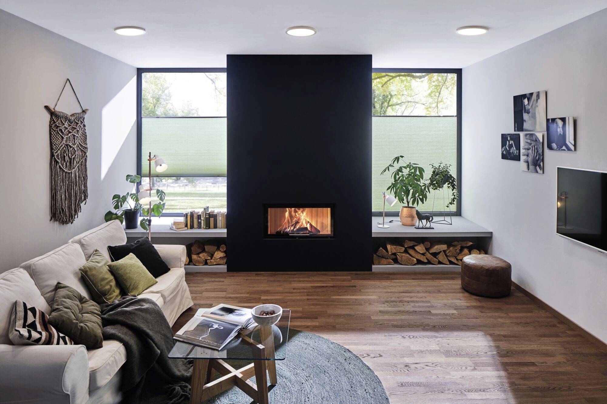 Helles Wohnzimmer mit Sofa, Kamin und Pflanzen. Große Fenster im Hintergrund. Gemütliche Dekoration, moderner Stil.