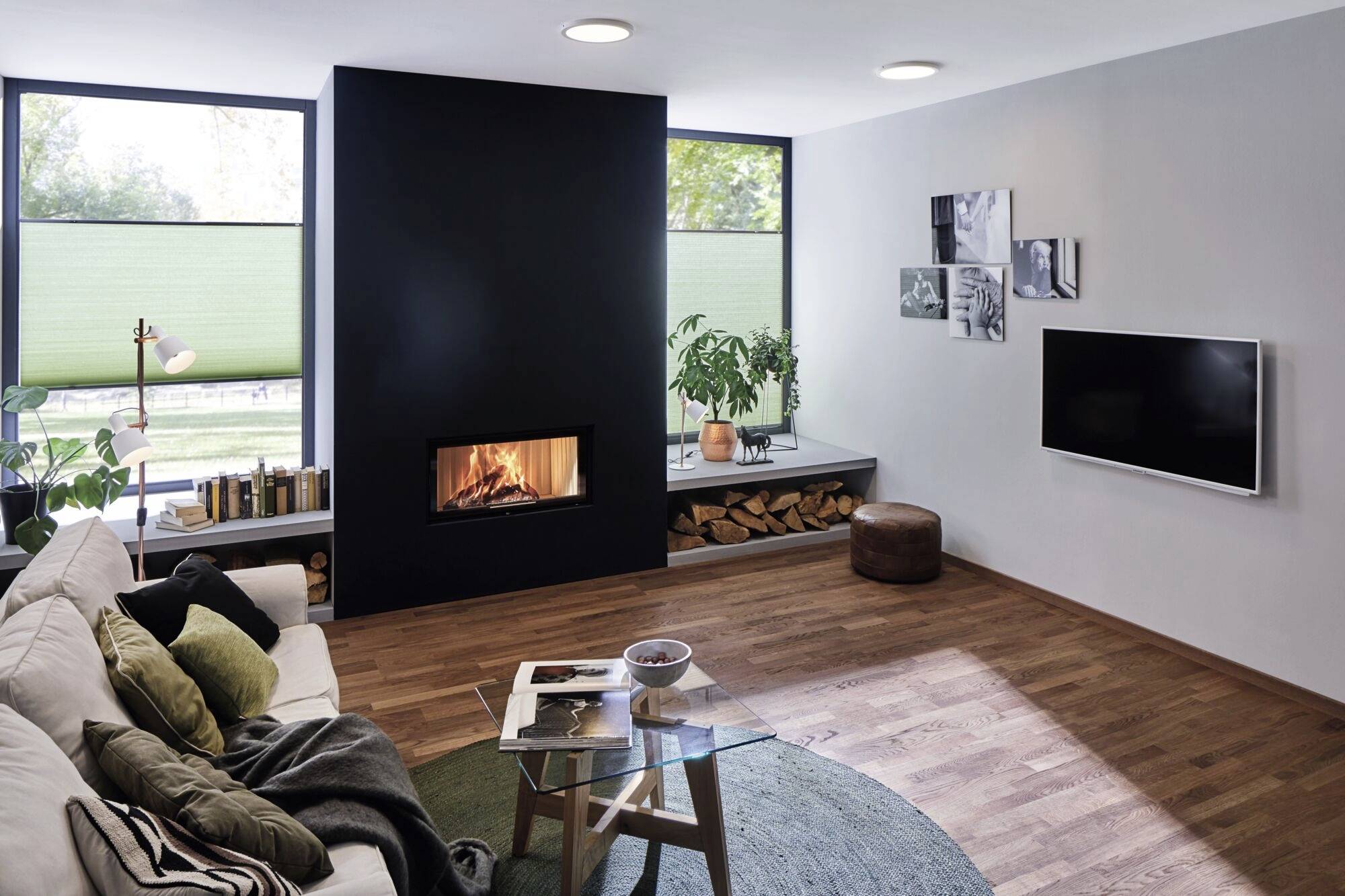 Offener Wohnzimmerbereich mit Kamin in modernem Design. Gemütliche Sitzecke, Bücherregal, Pflanzen und ein Flachbildfernseher an der Wand.