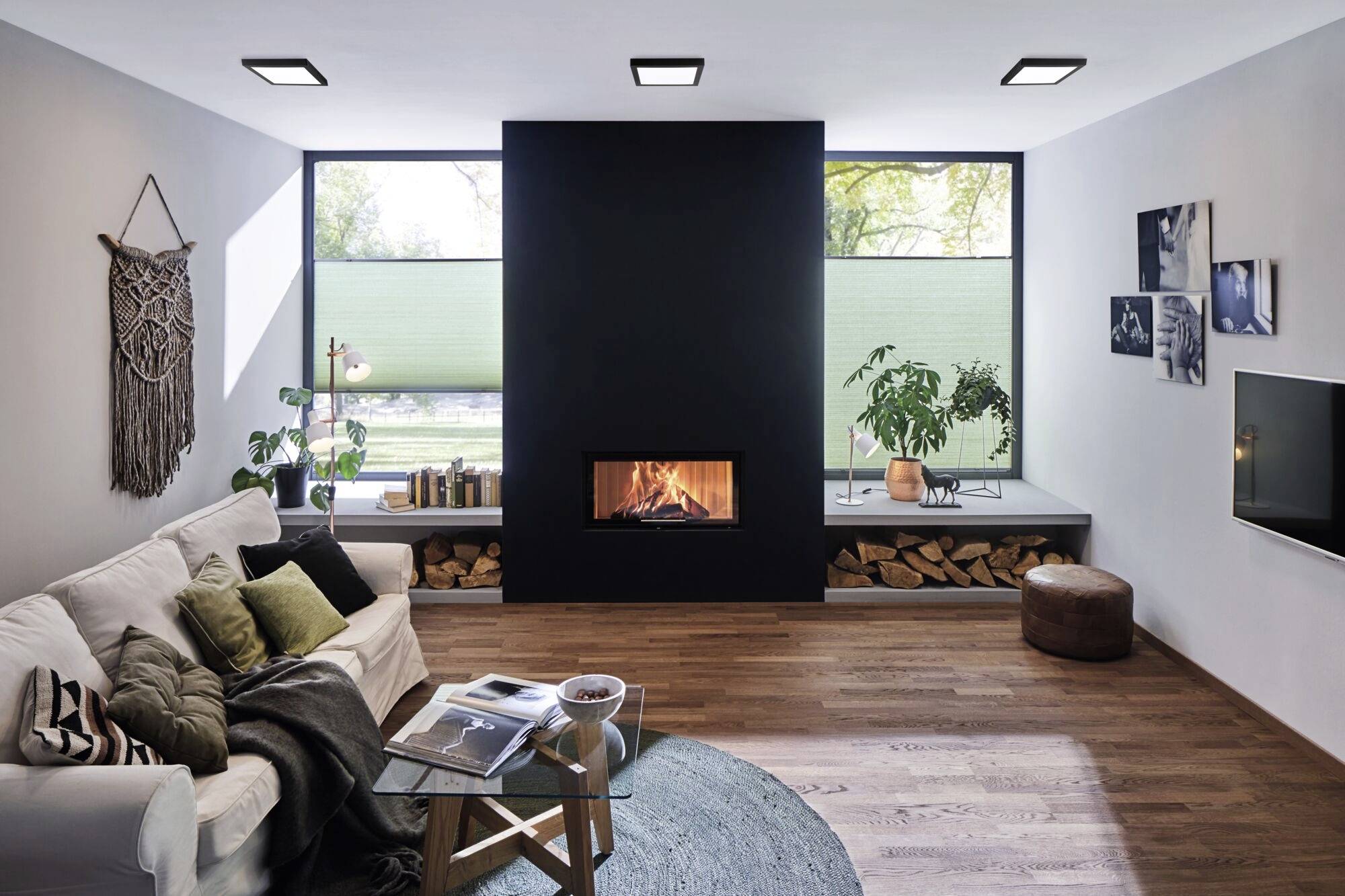 Modernes Wohnzimmer mit einem schwarzen Kamin im Zentrum, umgeben von Holzstapeln. Weiße Couch, Pflanzen und helle Fenster vervollständigen den Raum.