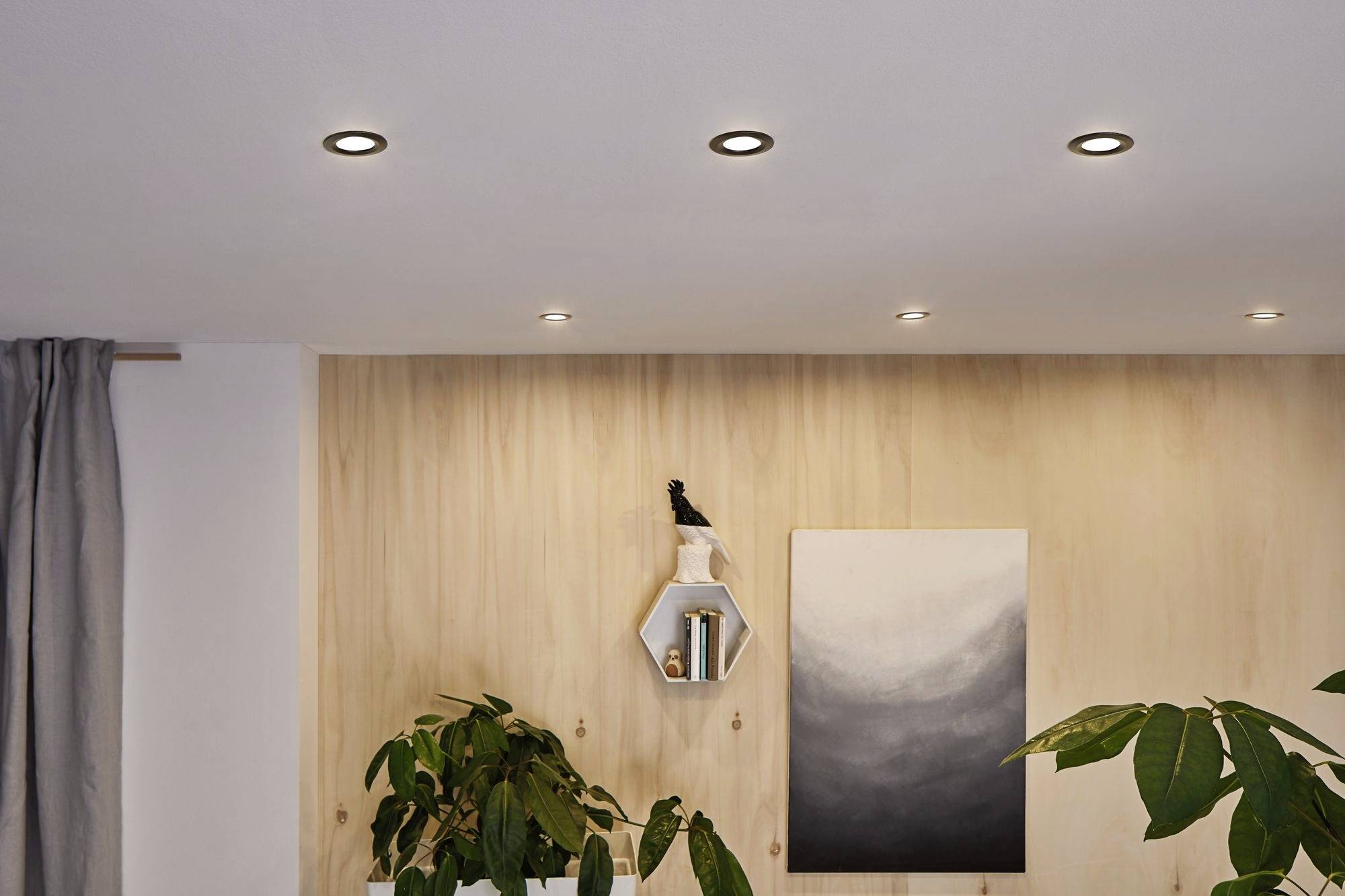 Ein Wohnzimmer mit Holzwand, hängendem Kunstwerk und Zimmerpflanzen. An der Decke sind sieben kleine, runde Einbaustrahler sichtbar.