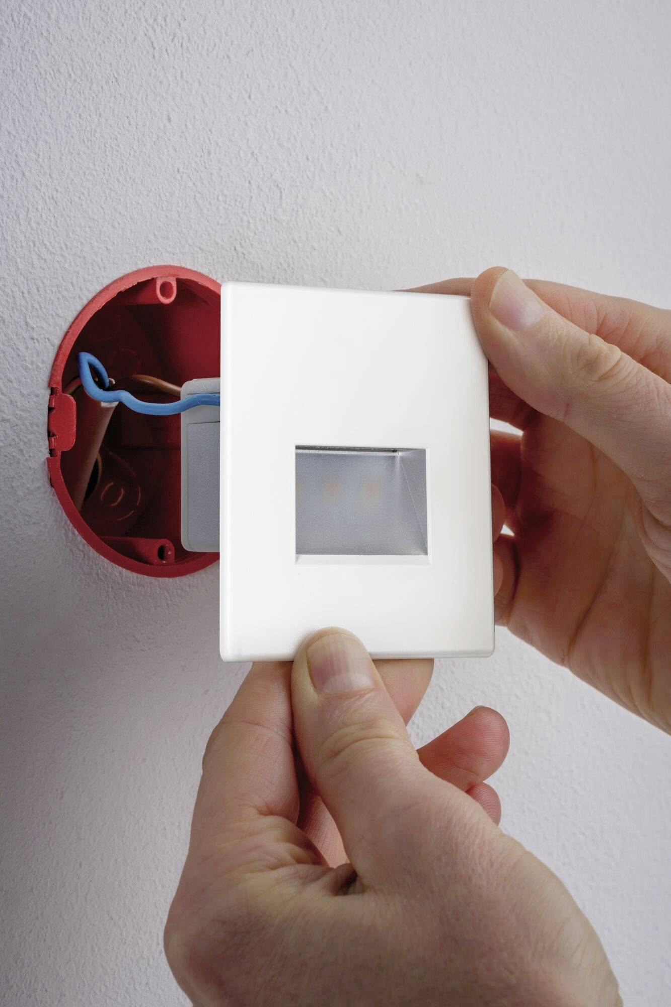 Nahaufnahme von Händen, die eine Abdeckplatte auf eine elektrische Steckdose an der Wand montieren. Blauer und brauner Draht sichtbar.