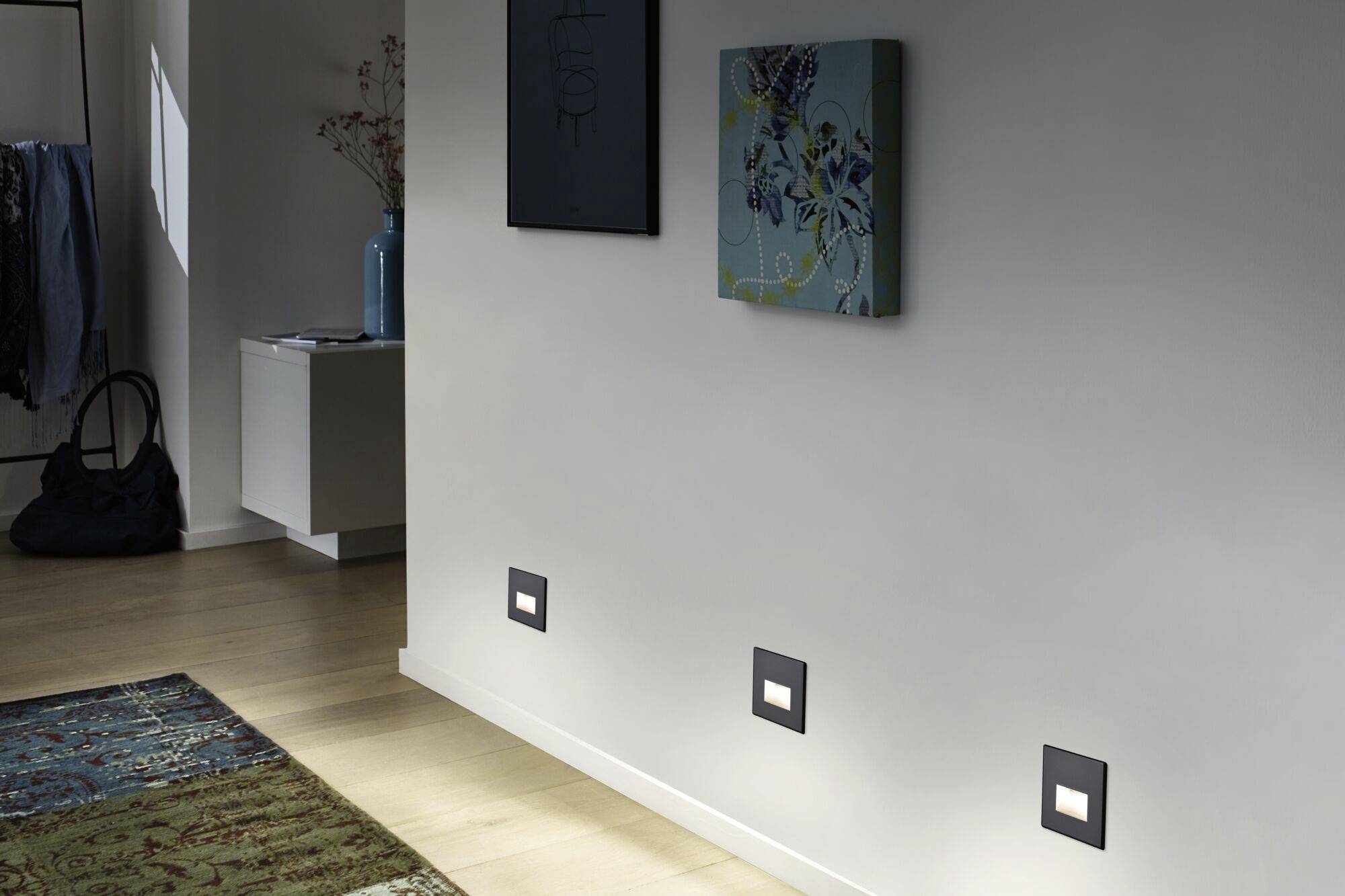 Dunkler Flur mit moderner Beleuchtung an der Wand und zwei Wandbildern. Teppich am Holzfußboden, dimme Lichtstimmung.