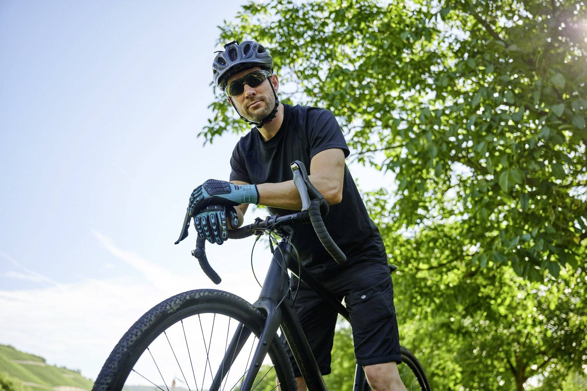 Ein Mann in Sportkleidung posiert mit einem Rennrad im Grünen, bei sonnigem Wetter. Er trägt einen Helm und Sonnenbrille.