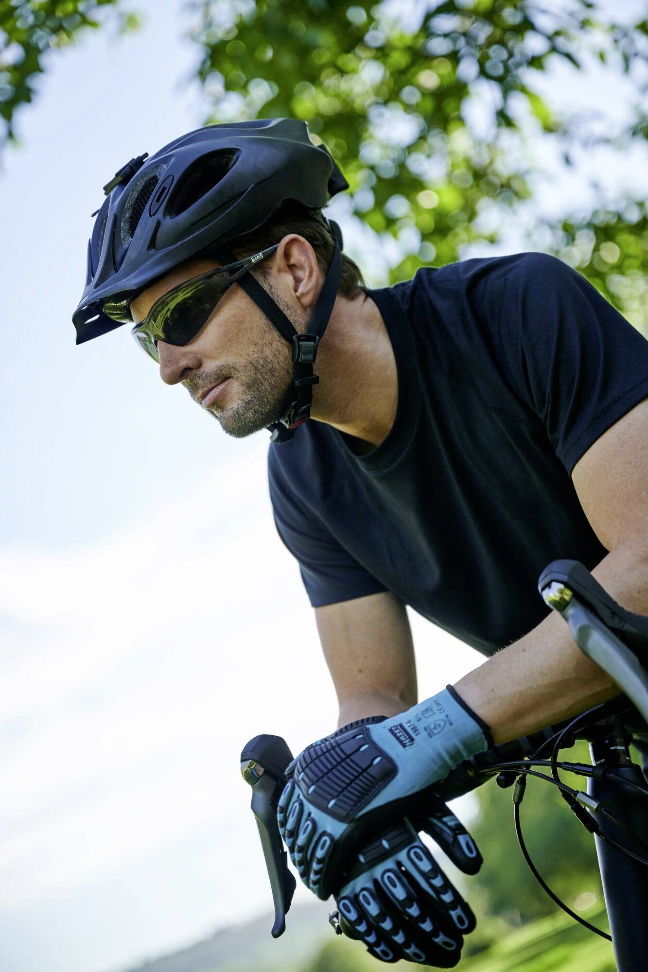 Ein Mann mit Helm und Sonnenbrille fährt Fahrrad in einer grünen Landschaft, leichter Himmel. Er trägt ein schwarzes Shirt und Handschuhe.