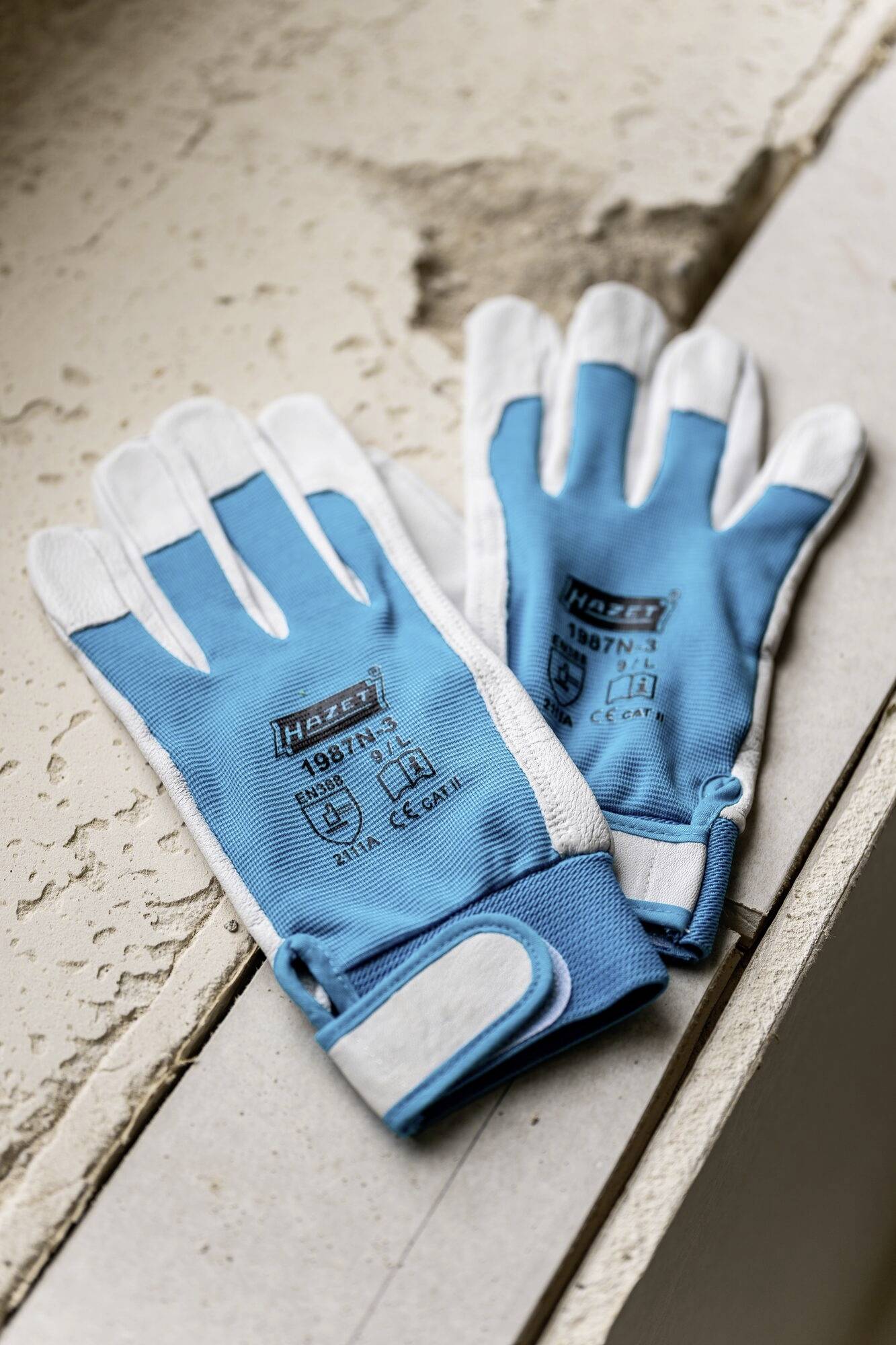 Zwei blaue Arbeitshandschuhe mit der Aufschrift 'Hazet 1987N-9' liegen auf einer groben Betonoberfläche.