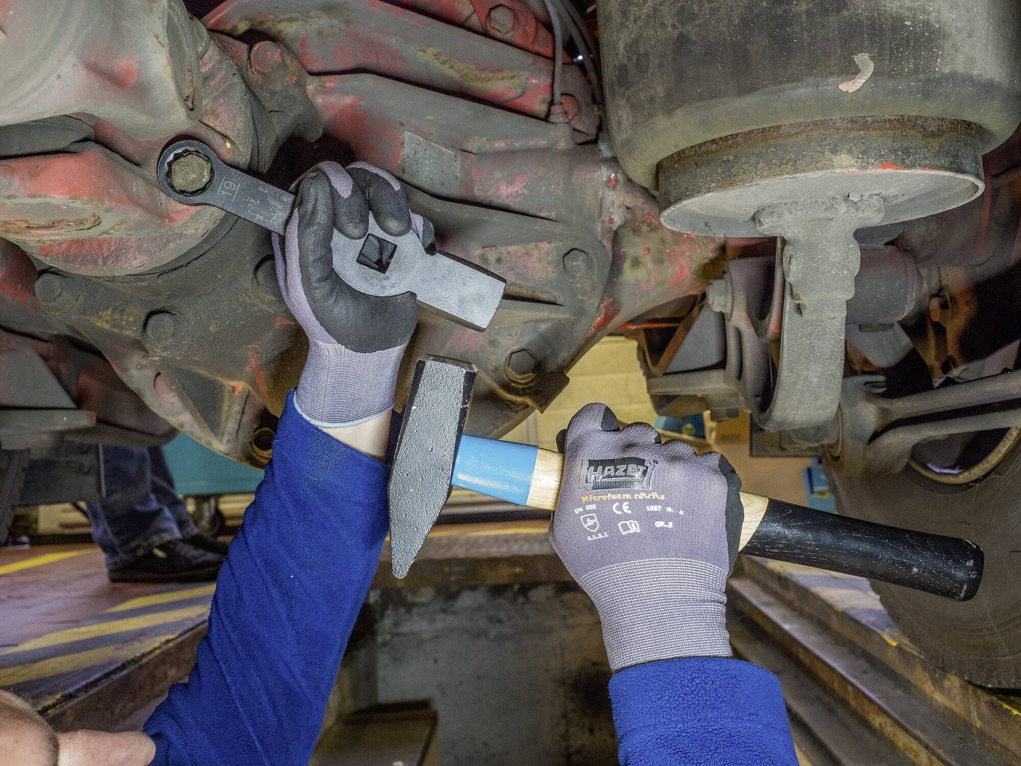 Mechaniker repariert Unterseite eines Fahrzeugs in einer Werkstatt, hält Schraubenschlüssel und Hammer.