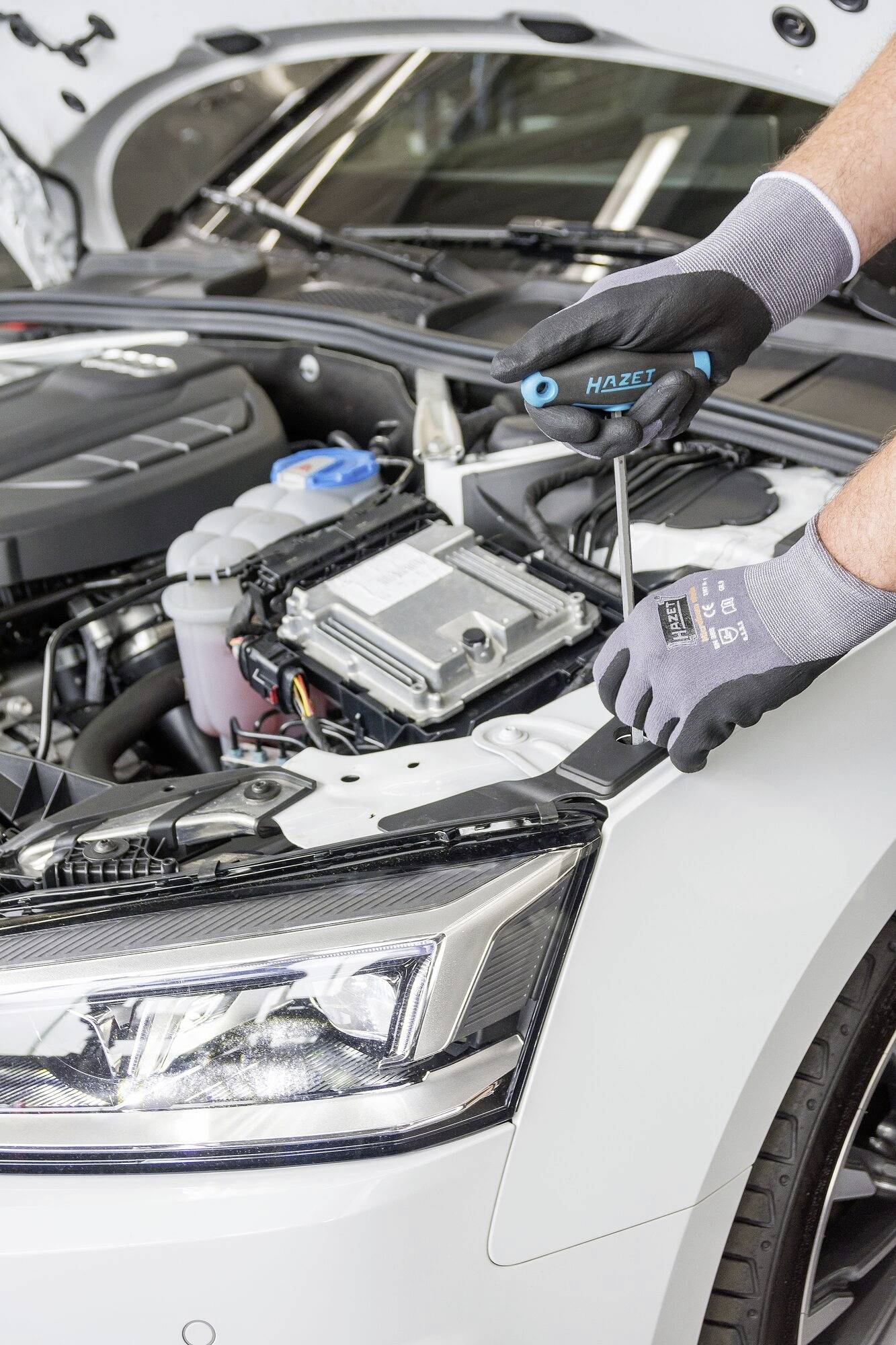 Ein Mechaniker arbeitet mit einem Schraubenzieher am Motorraum eines Autos, das mit geöffneter Motorhaube in einer Werkstatt steht.
