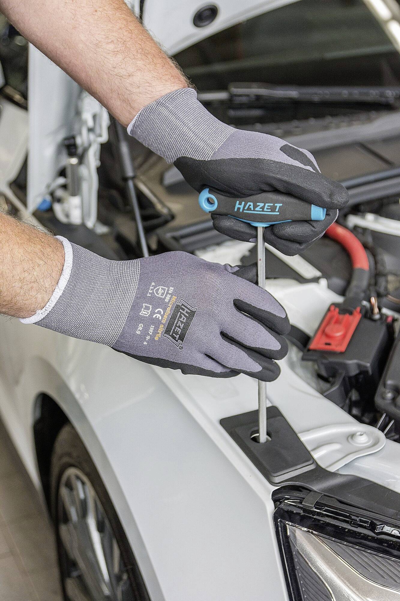 Eine Person repariert mit einem Schraubendreher vor der geöffneten Motorhaube eines Autos. Sie trägt graue Handschuhe.