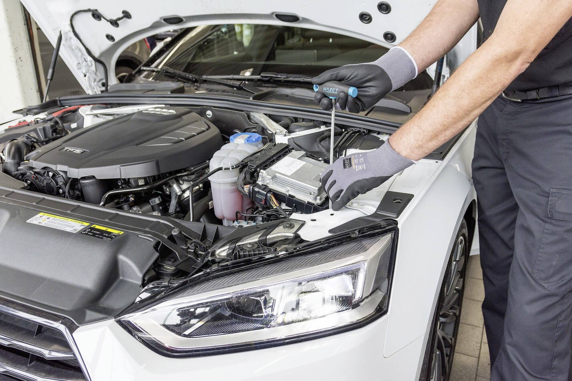 Mechaniker arbeitet am Motor eines weißen Autos mit offenem Motorraum und benutzt einen Schraubendreher.