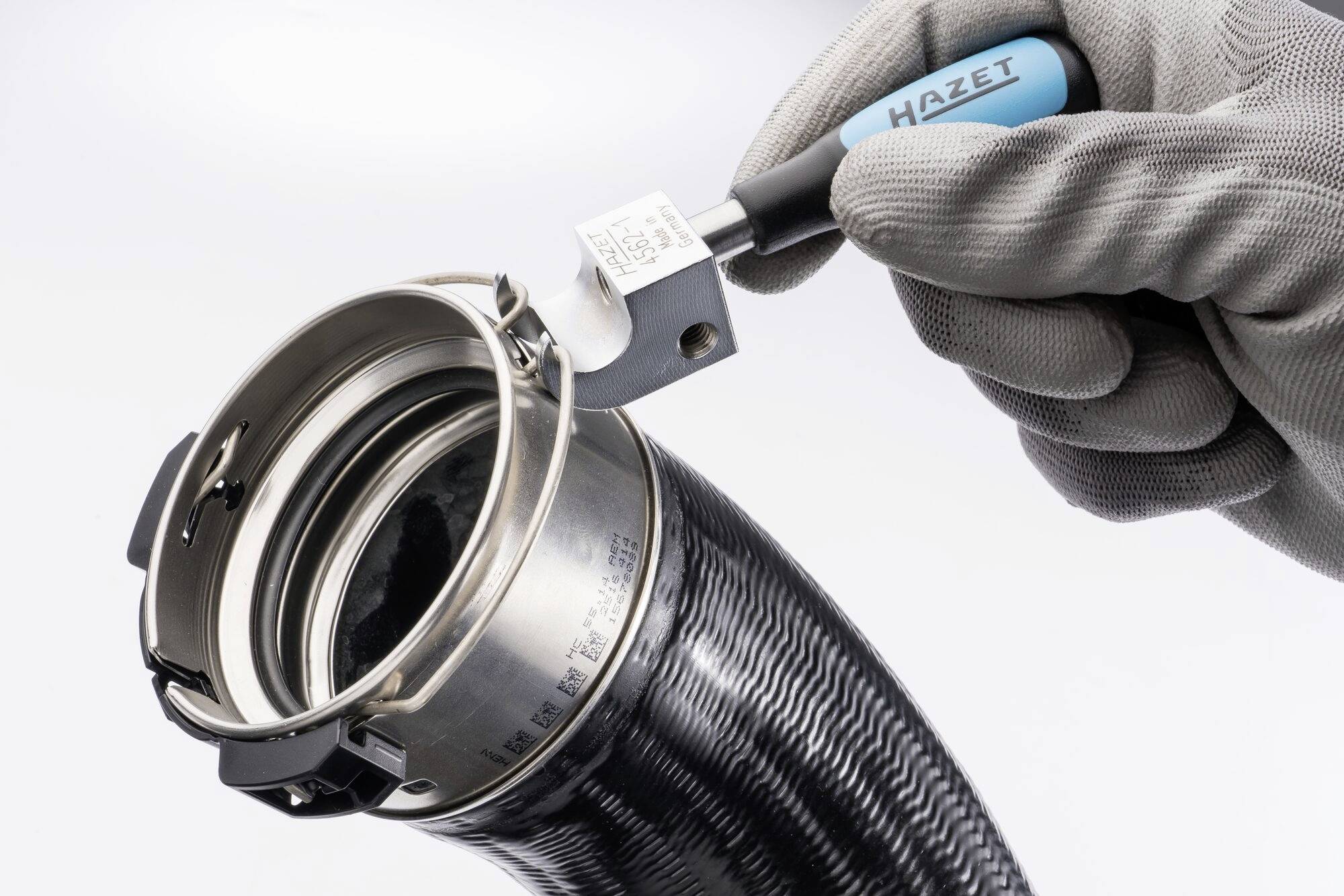 Hand mit Handschuh hält ein blau-schwarzes Werkzeug von 'HAZET' an einer metallenen Schlauchschelle, die ein Rohrstück umschließt.