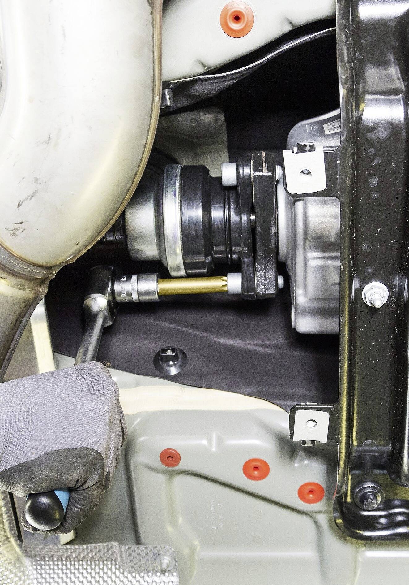 Eine Person zieht mit einem Schraubenschlüssel an einem Riegel im Automechanikbereich, nahe einem Auspuffrohr und einer schwarzen Metallhalterung.