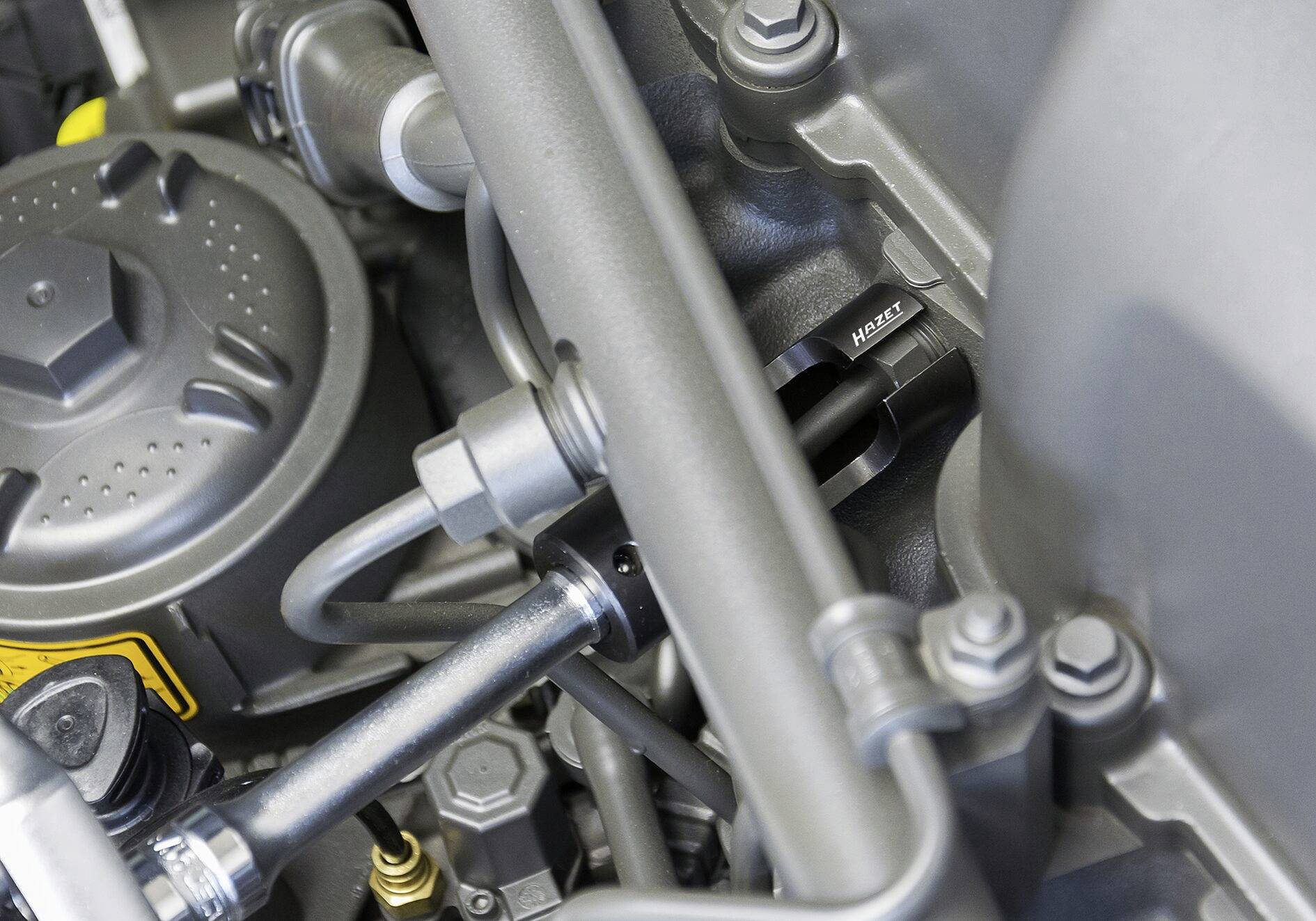 Blick auf den Motorraum eines Fahrzeugs mit Fokussierung auf verschiedene mechanische Teile und Schläuche.