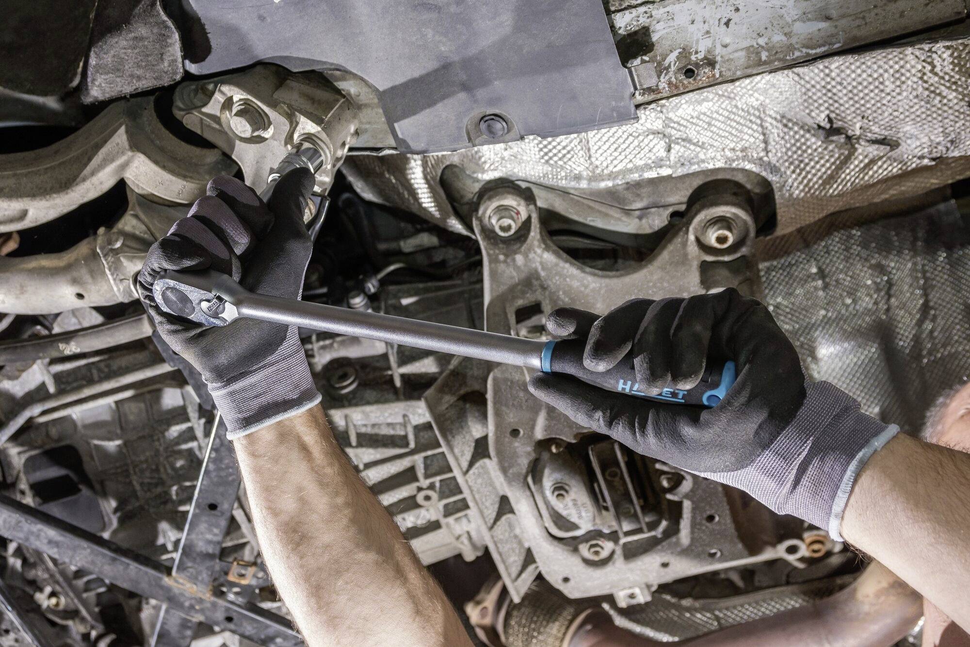 Mechaniker zieht Schraube an Autochassis an, Nahaufnahme. Werkzeug mit Drehmomentschlüssel, Arbeitshandschuhe, Metallstruktur erkennbar.