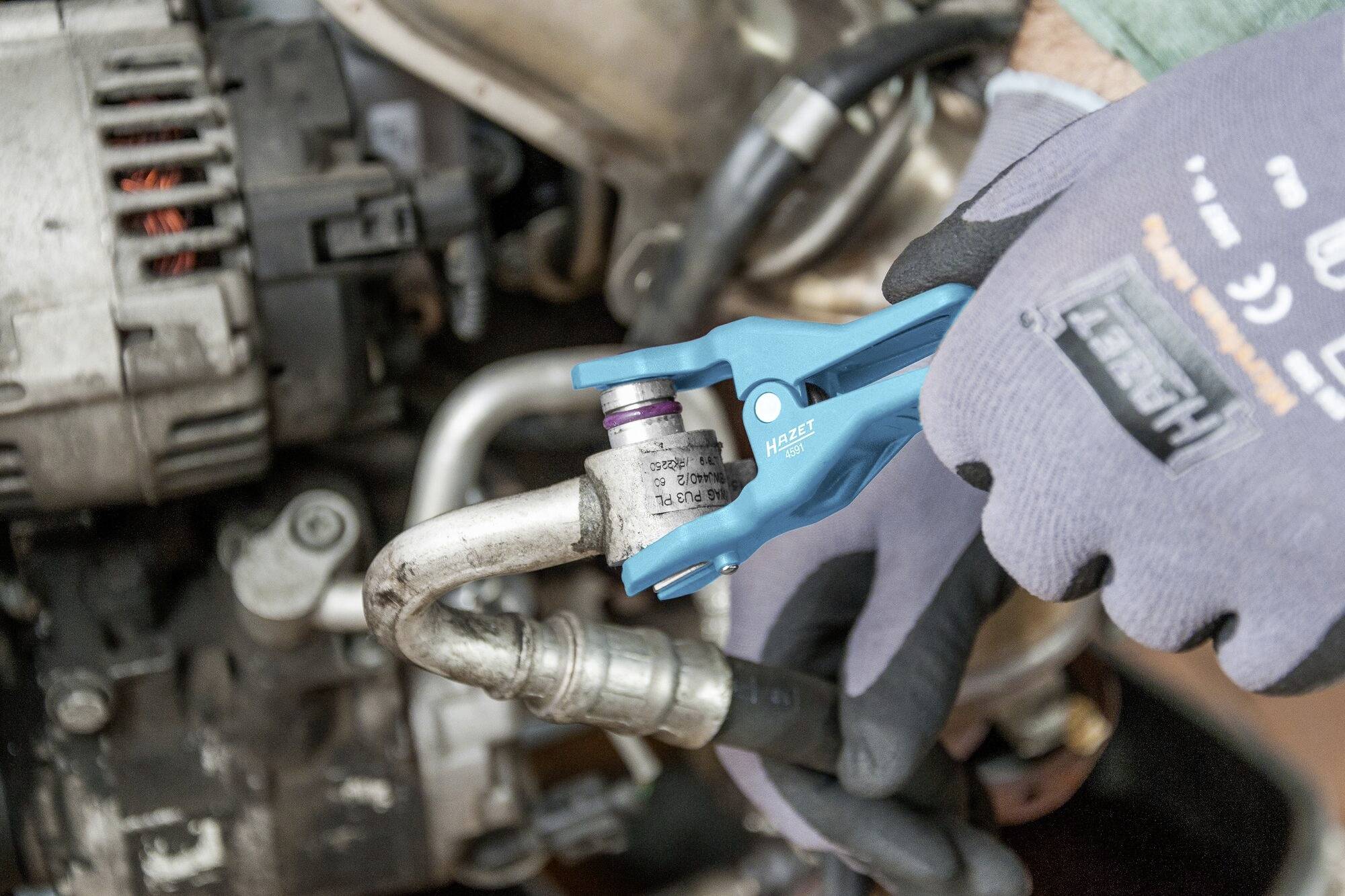 Eine Person mit Schutzhandschuhen hält eine blaue Zange und arbeitet an einer Metallverbindung in einem Automotor.