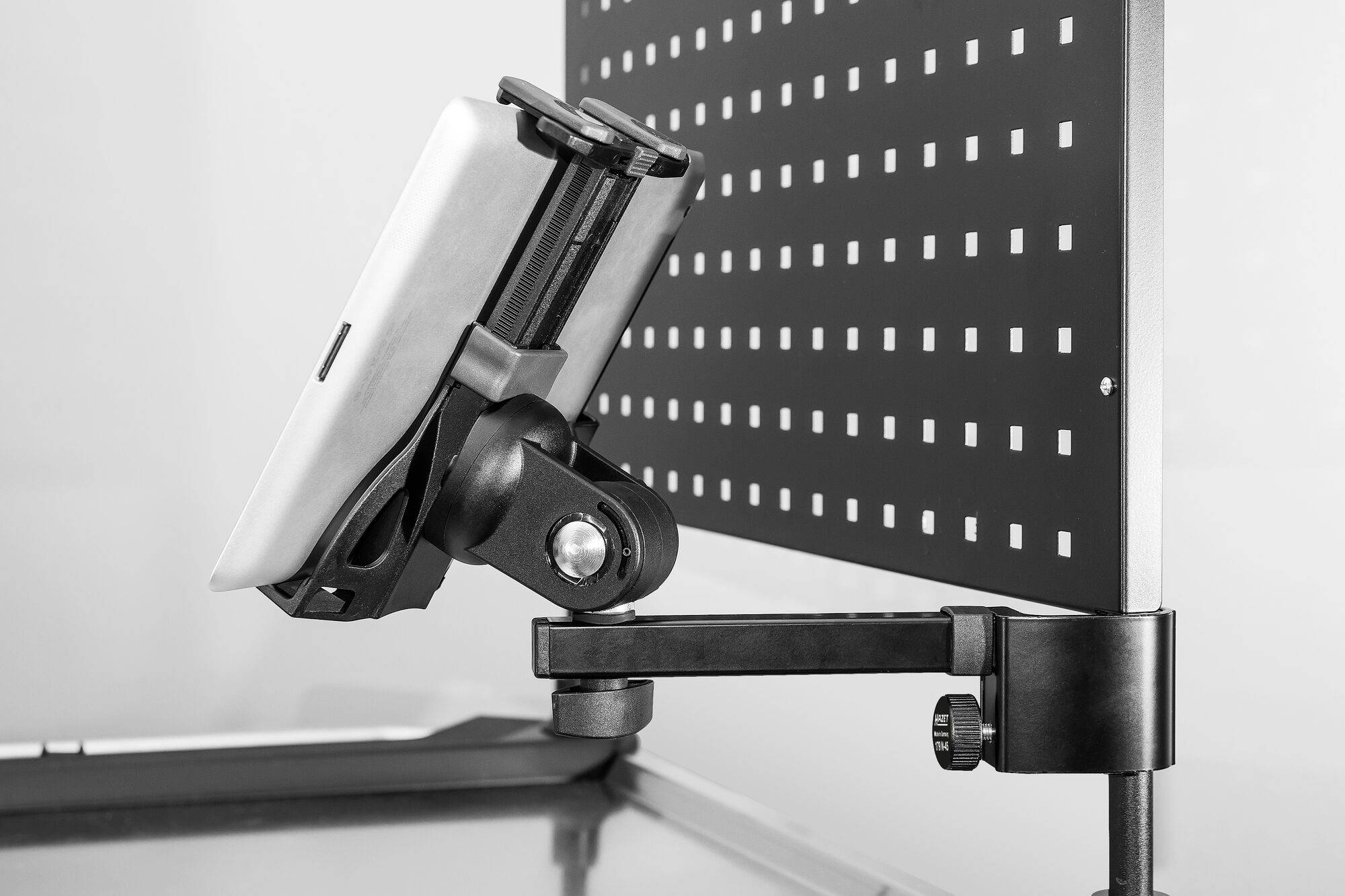 Ein Tablet ist in einem verstellbaren Halter an einer perforierten Tafel befestigt. Der Hintergrund ist unscharf.