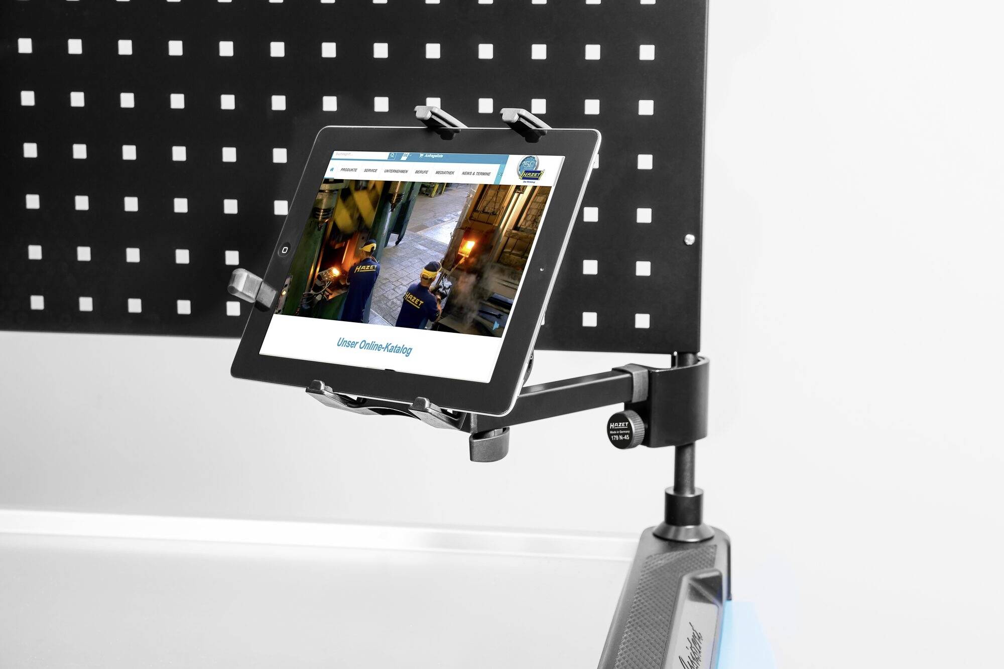 Ein Tablet in einer Halterung, zeigt eine Webseite mit dem Titel 'Unser Online-Katalog'. Hintergrund: Lochplatte aus Metall.