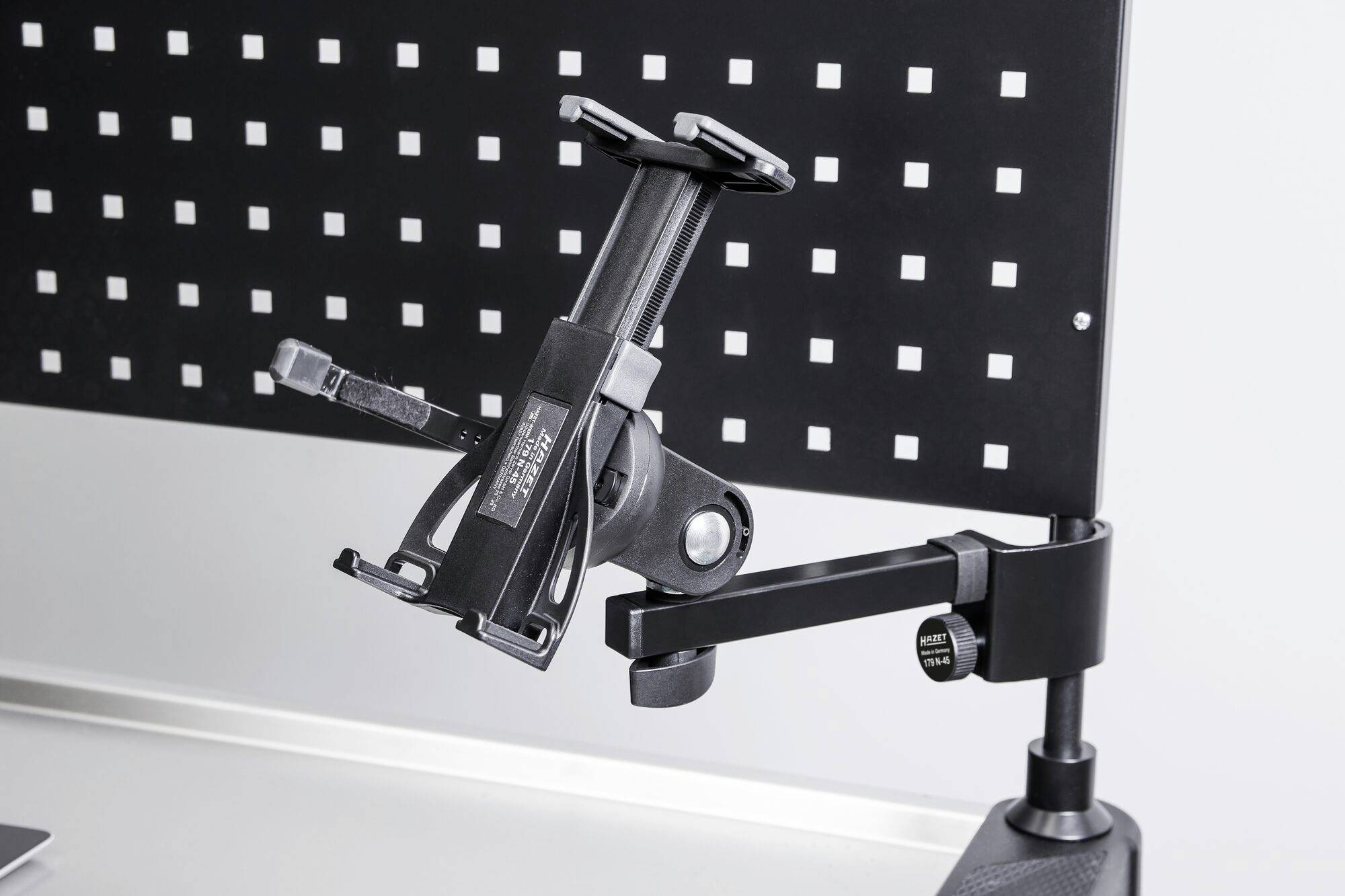 Ein schwarzer Tablet-Halter mit verstellbarem Arm ist an einer perforierten Wand befestigt und dient zur flexiblen Positionierung eines Geräts.