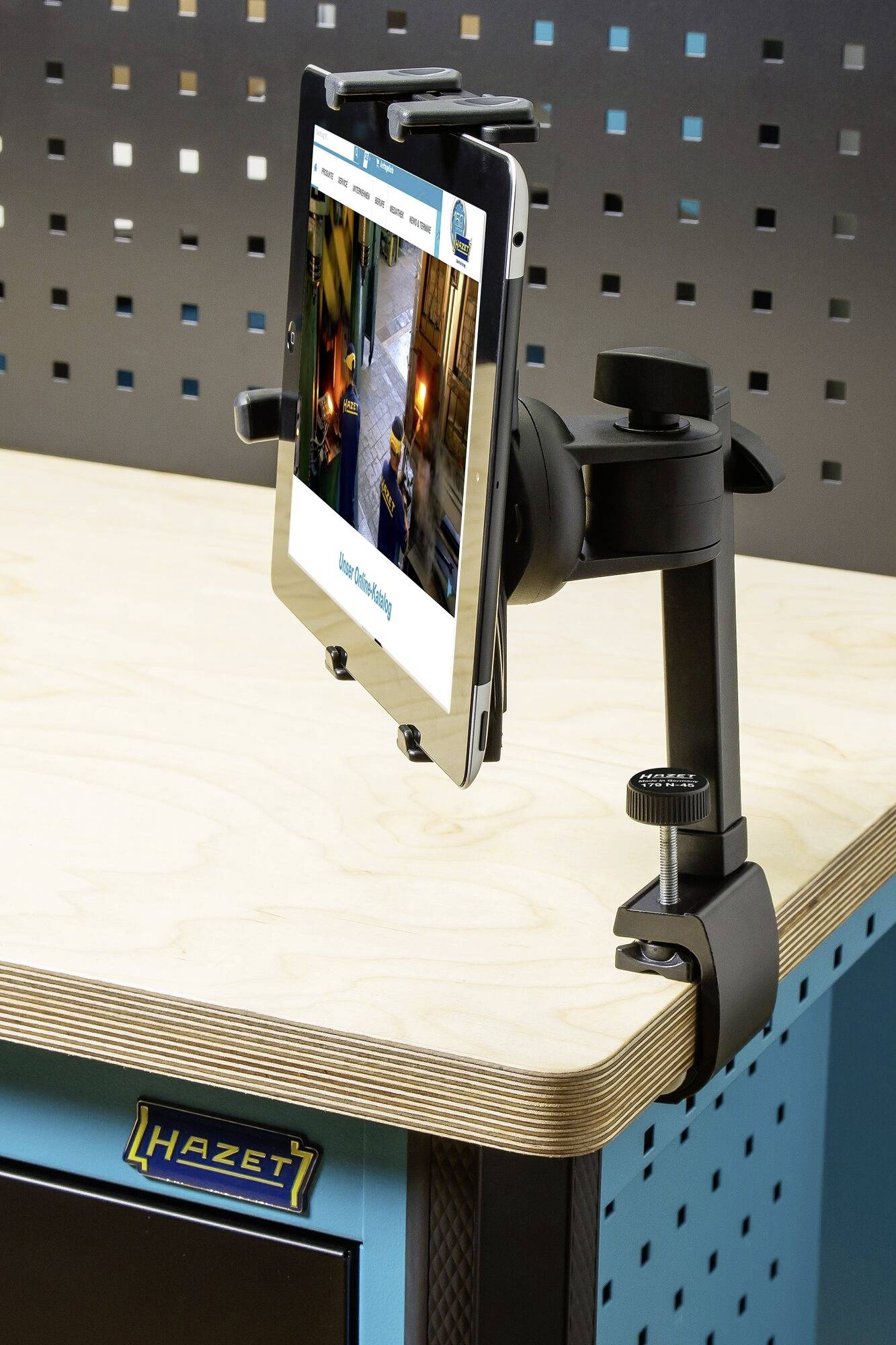 Eine Tablet-Halterung ist an einem Tisch befestigt. Sie zeigt ein Tablet mit einer offenen Webseite. Die Umgebung wirkt technisch modern.