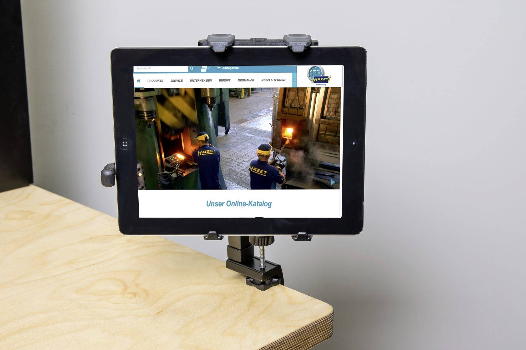 Ein Tablet mit einer geöffneten Webseite zeigt 'Unser Online-Katalog'. Es ist in einer Halterung auf einem Tisch befestigt.