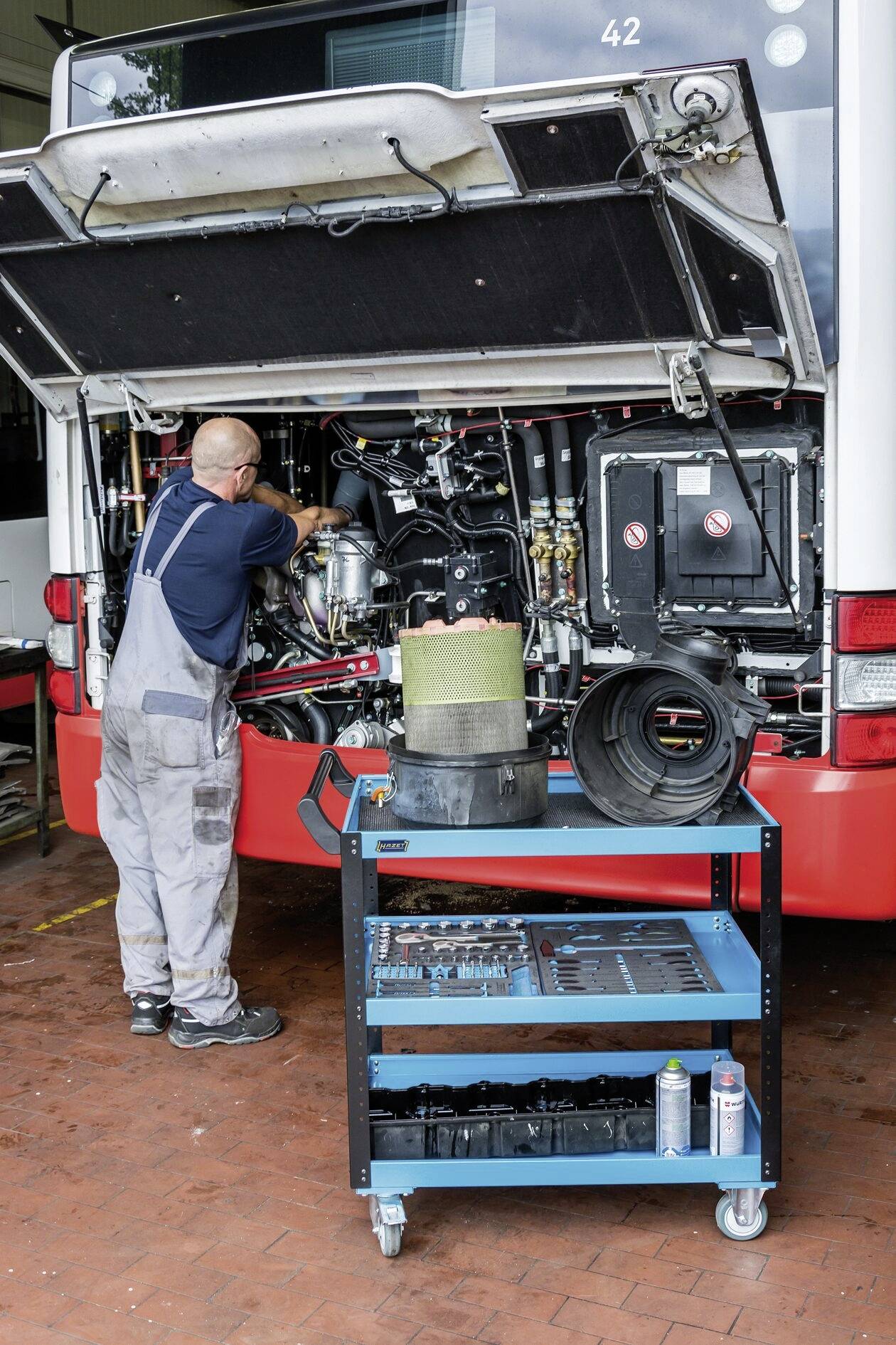 Mechaniker arbeitet am Motor eines Busses in einer Werkstatt. Ein Werkstattwagen mit Werkzeugen und Teilen steht im Vordergrund.